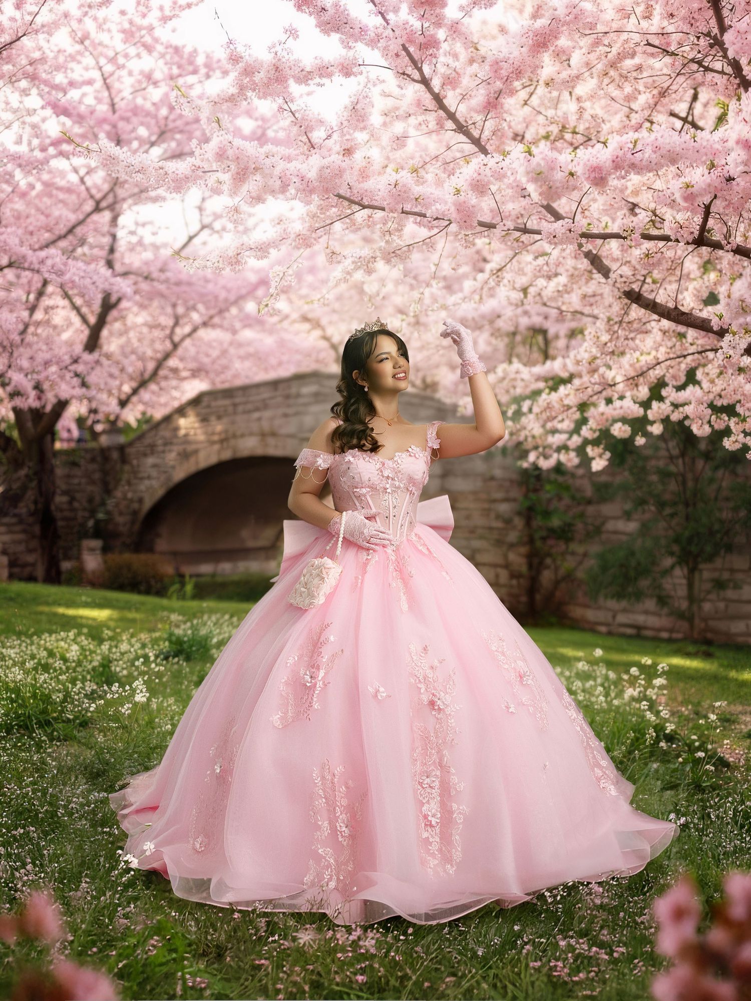 Young woman in pink ball gown poses under blooming cherry blossom trees near stone bridge.