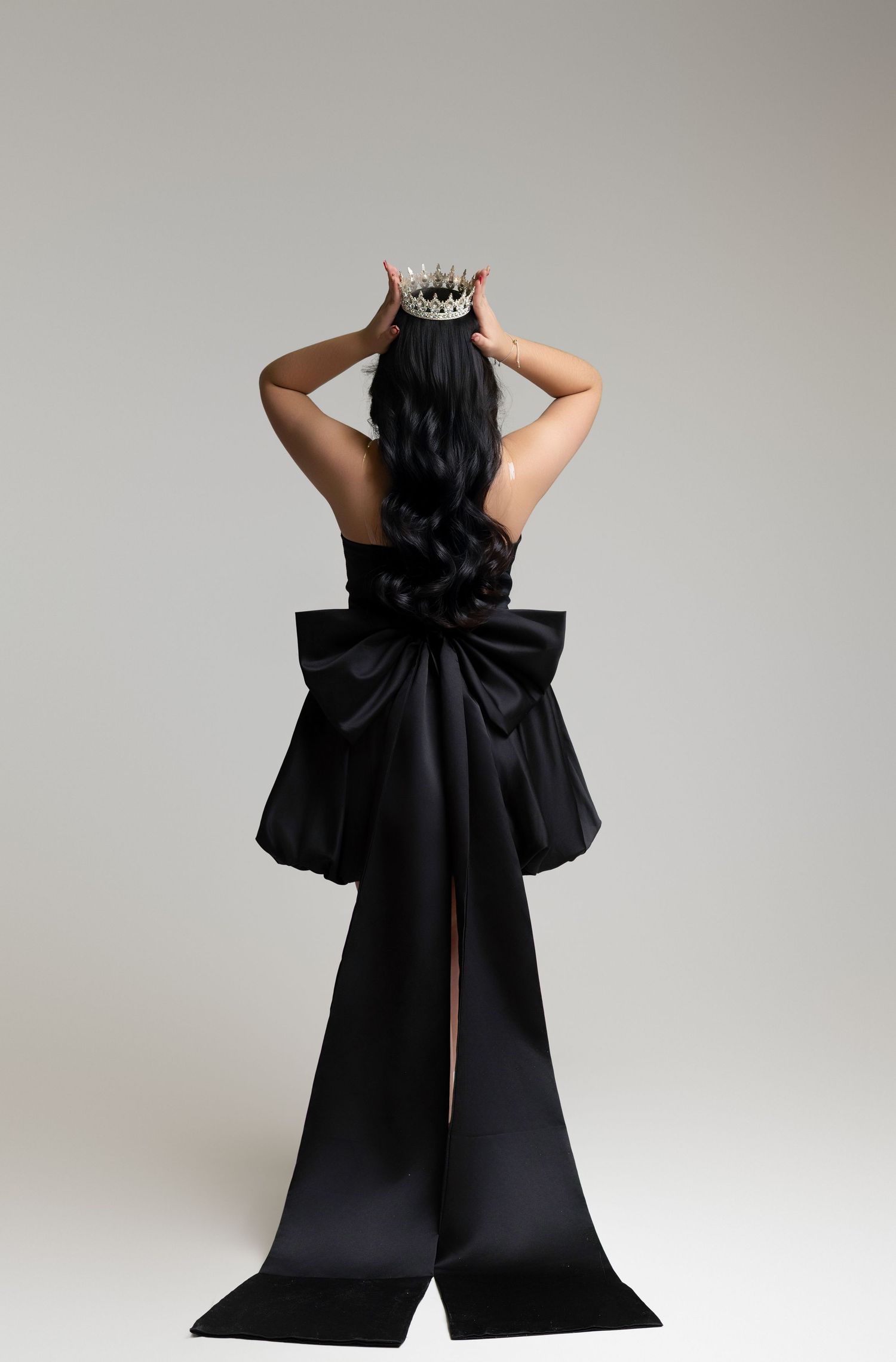 Woman in black satin gown with large bow, wearing crown, viewed from behind on white background.