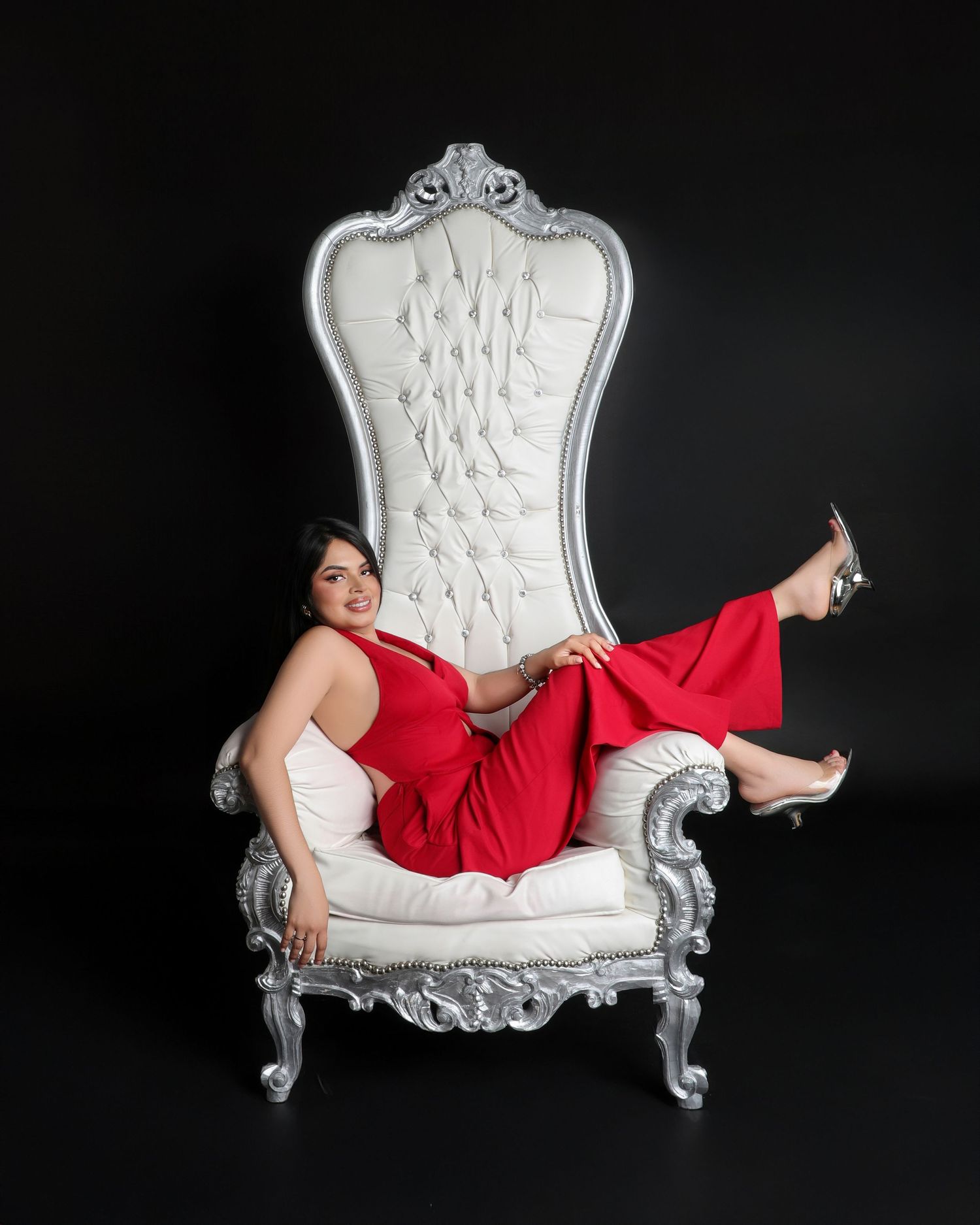 Woman in red dress posing elegantly on a silver tufted throne chair against a black background.