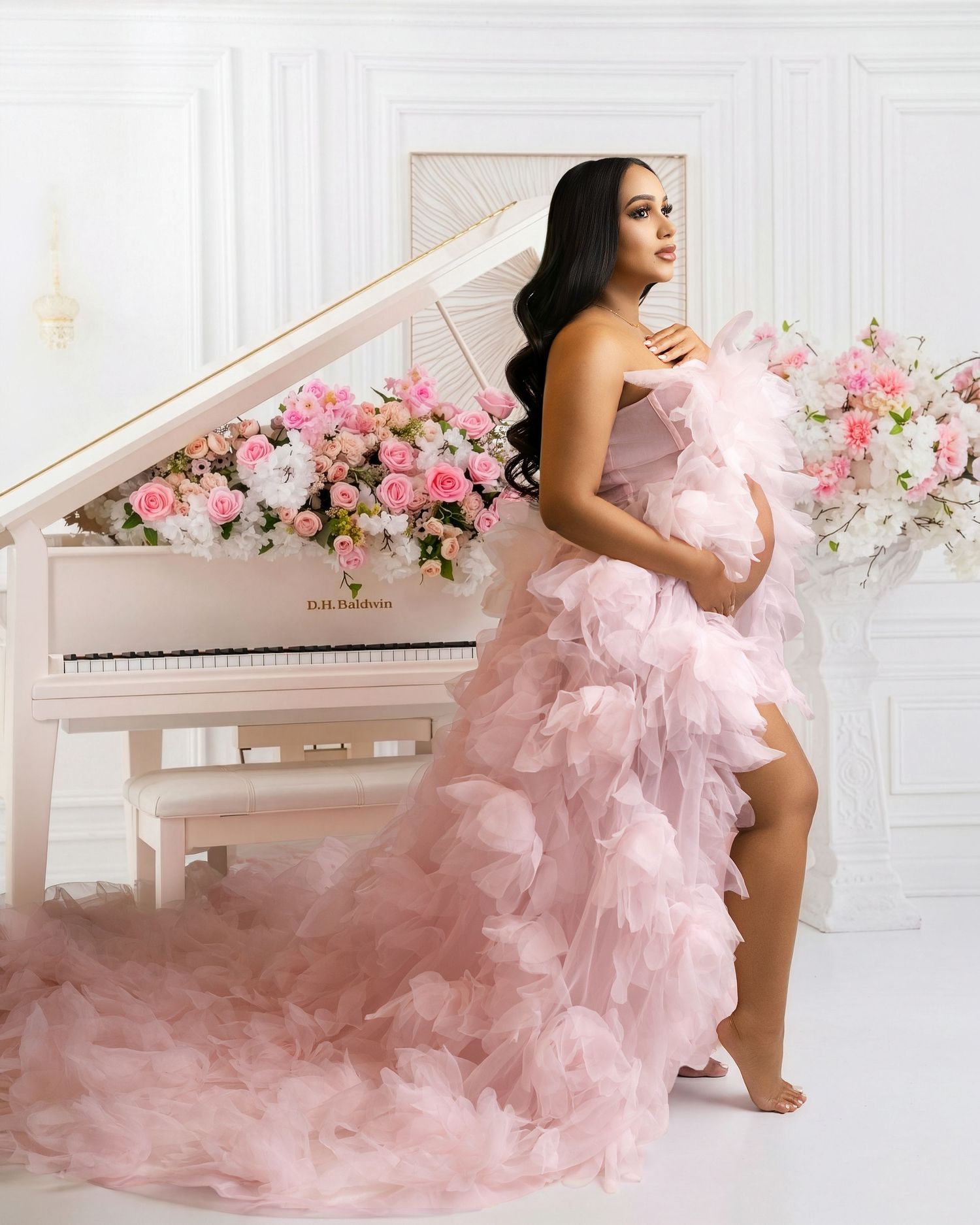 Pregnant woman in a pink ruffled tulle maternity gown posing beside a white grand piano with floral arrangements.