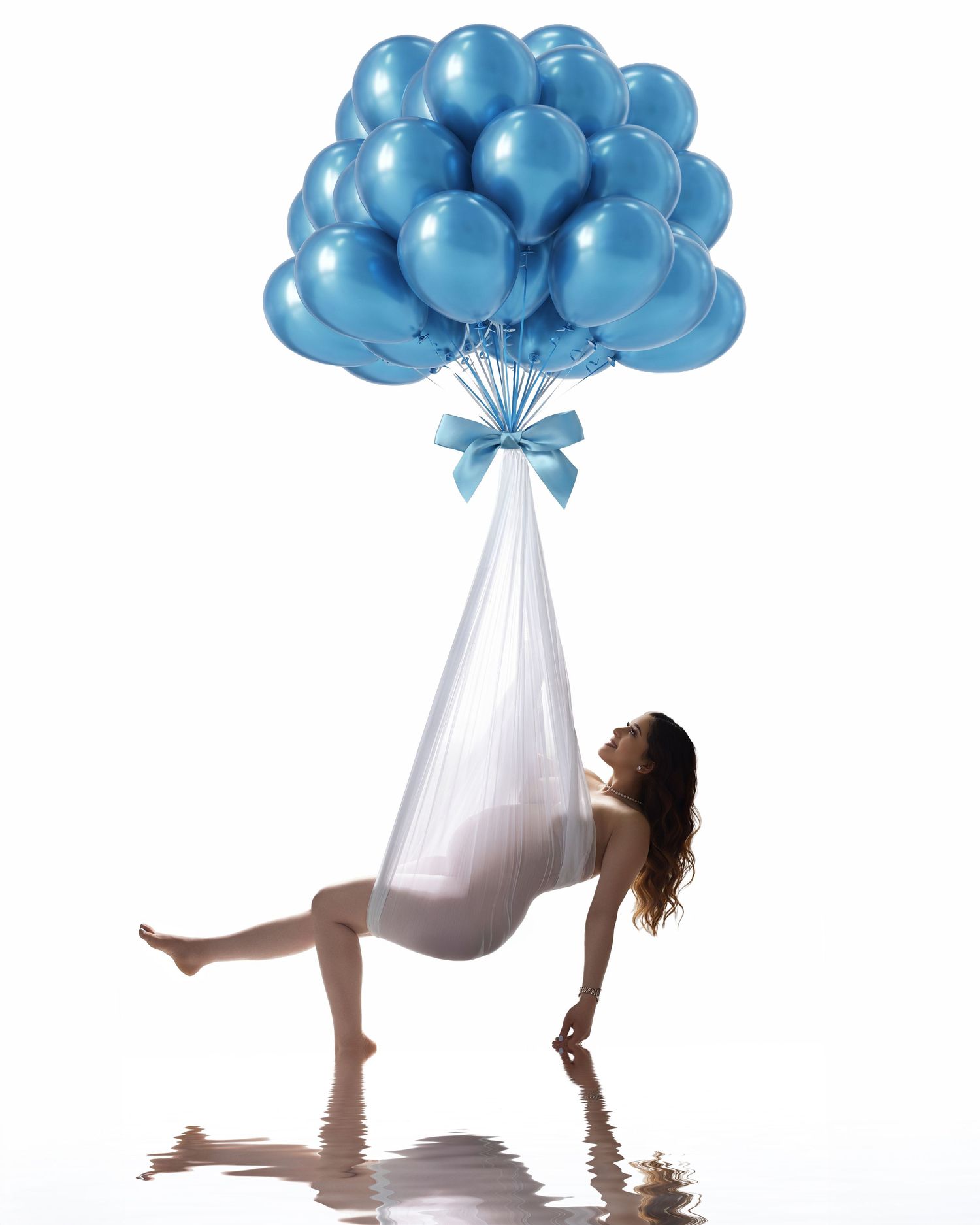 Pregnant woman in white dress lifted by blue balloons tied with ribbon, reflected on water surface.