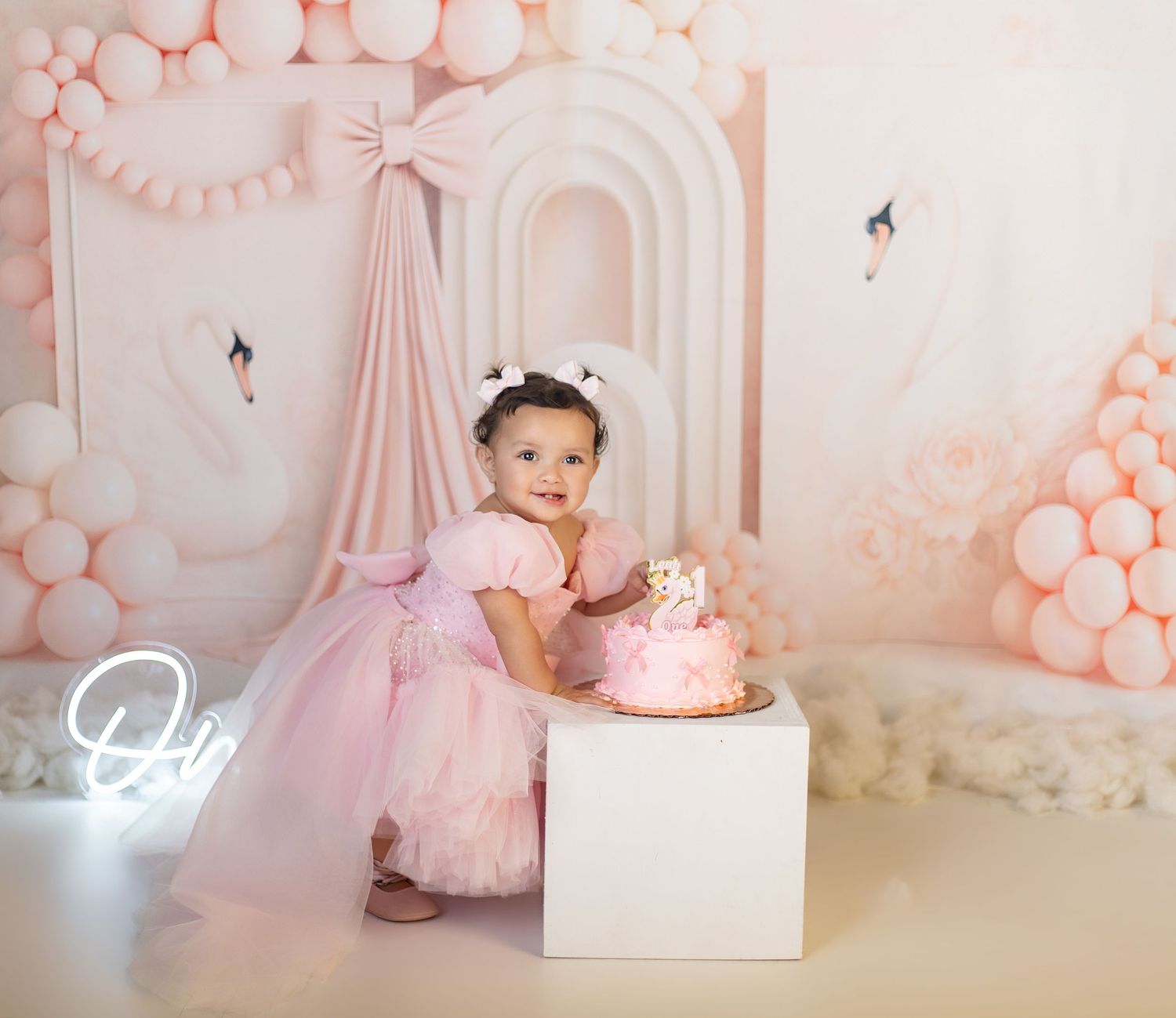 Toddler girl in pink tutu dress celebrates first birthday with cake amid pink balloon arch backdrop.