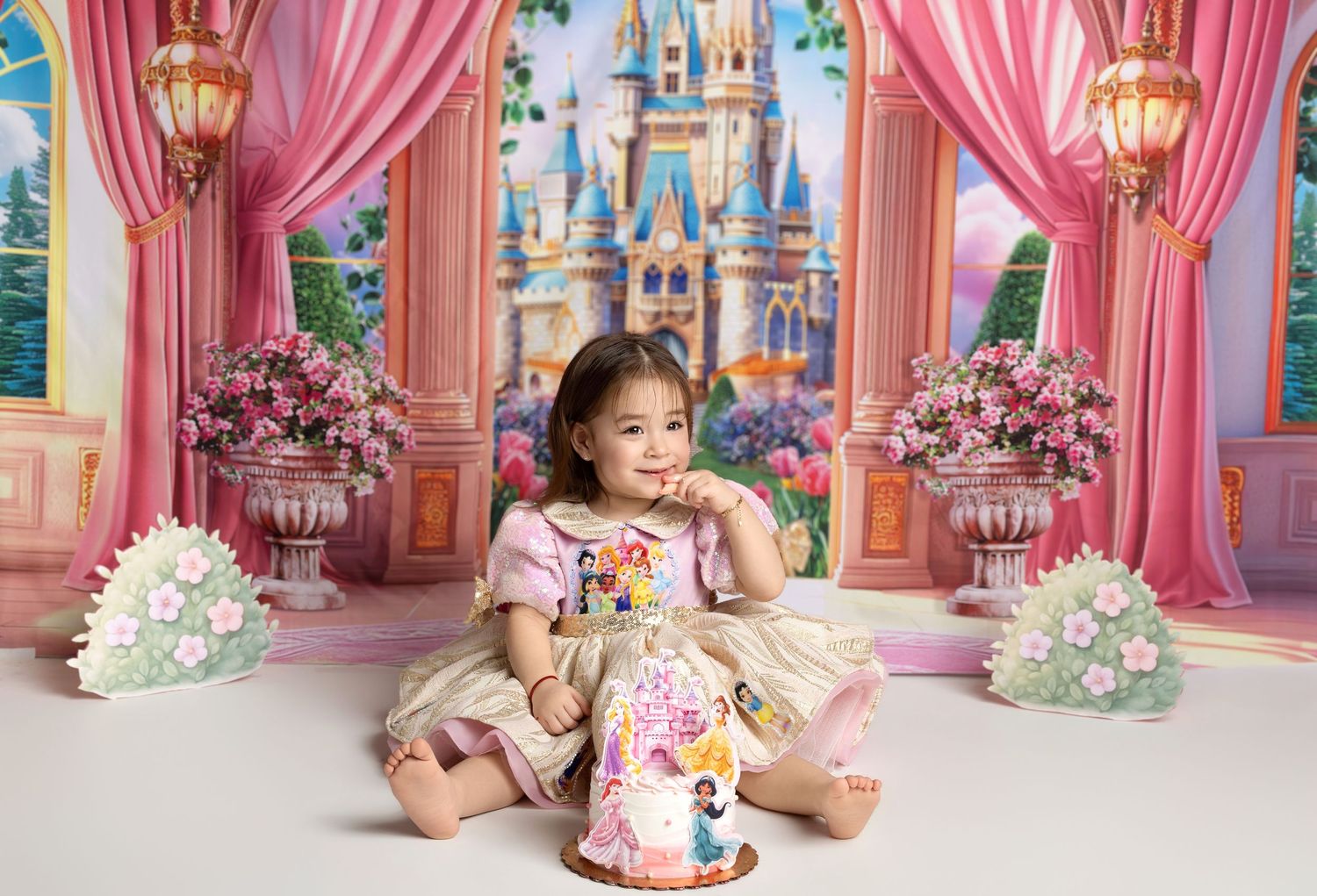 A toddler girl in a princess dress sits beside a Disney castle cake at a pink royal castle birthday photo shoot.