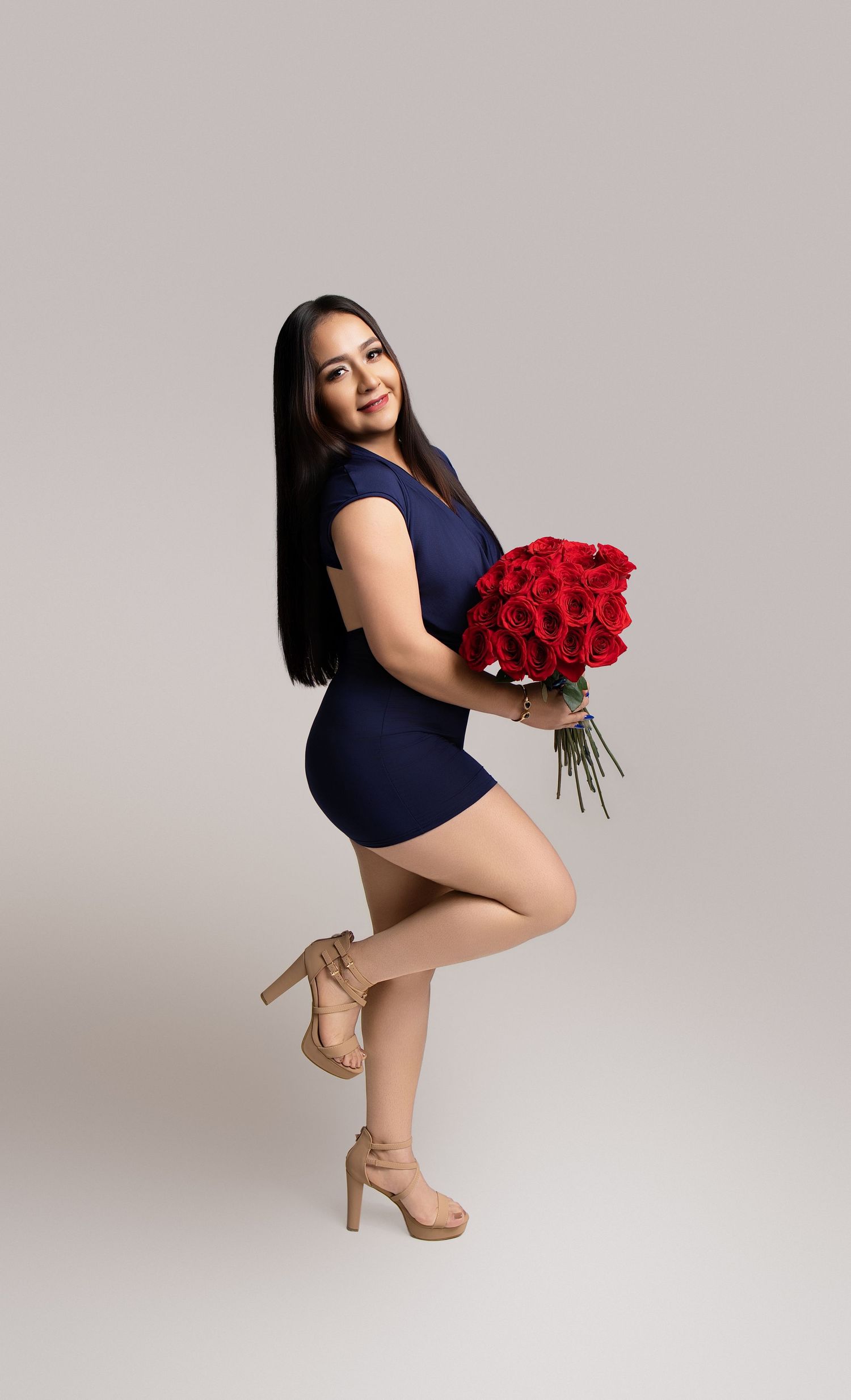 Woman in navy blue mini dress holding red roses bouquet, posing in heels against gray studio background.