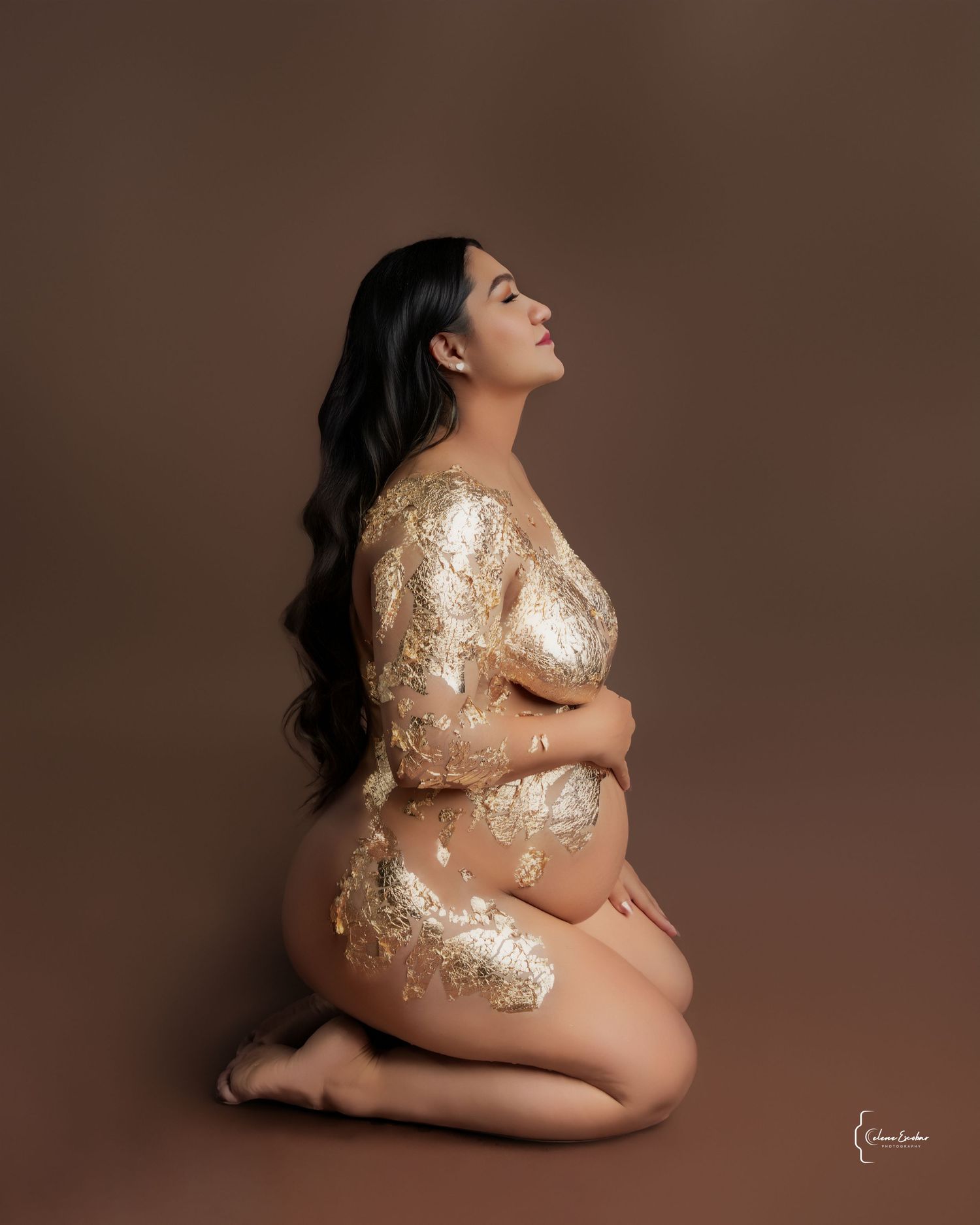 Elegant maternity portrait of a pregnant woman covered in gold foil on a brown background.