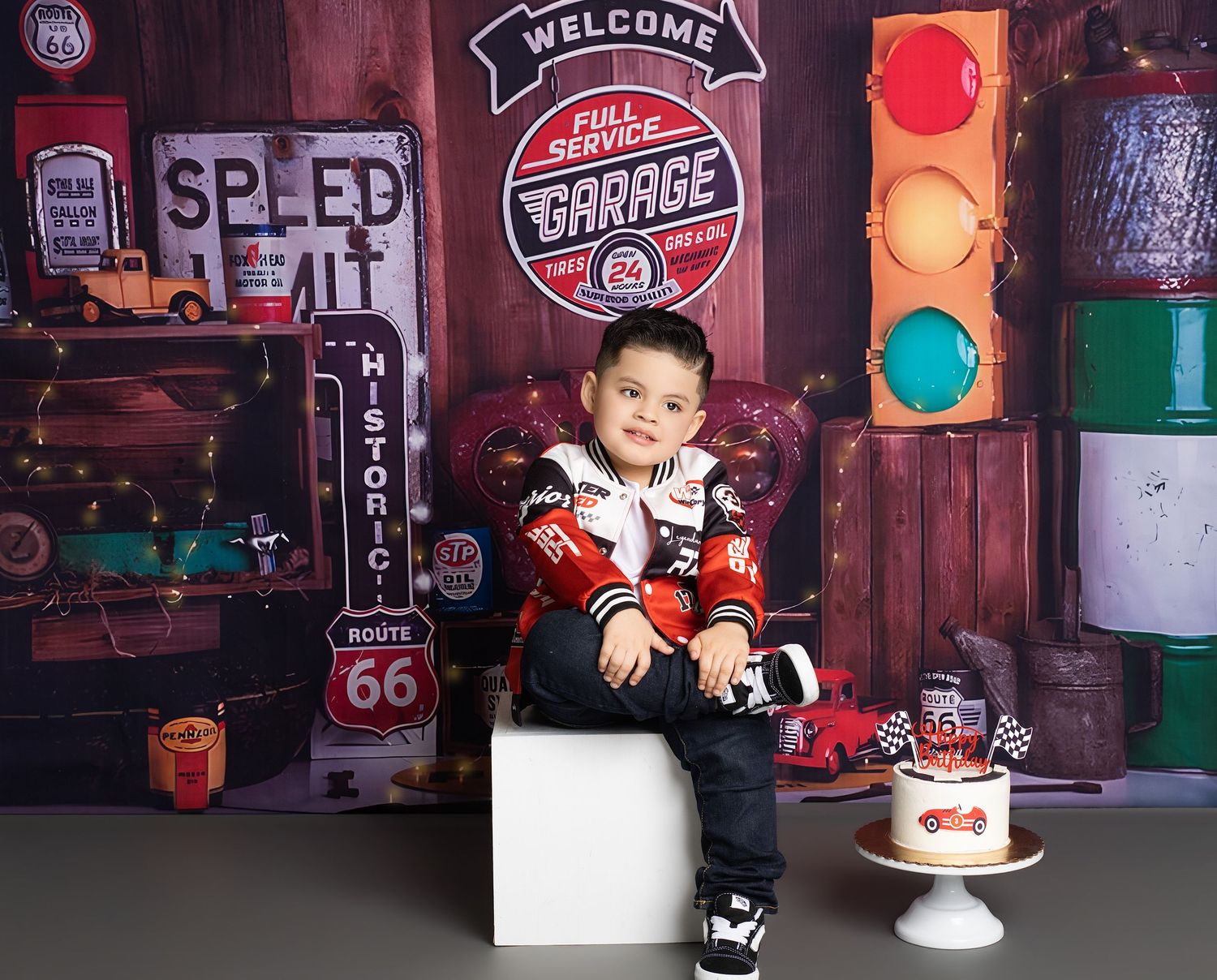 Young boy sits on white cube by Cars-themed birthday cake, Route 66 garage backdrop with traffic light decor.
