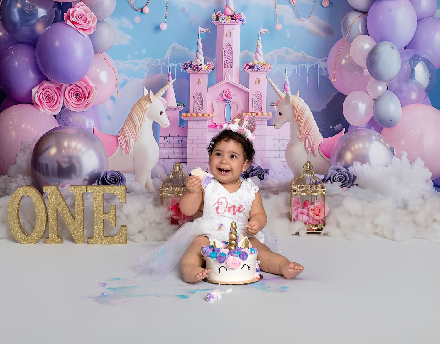 Baby girl celebrates first birthday with unicorn cake, balloon backdrop, and castle decorations.
