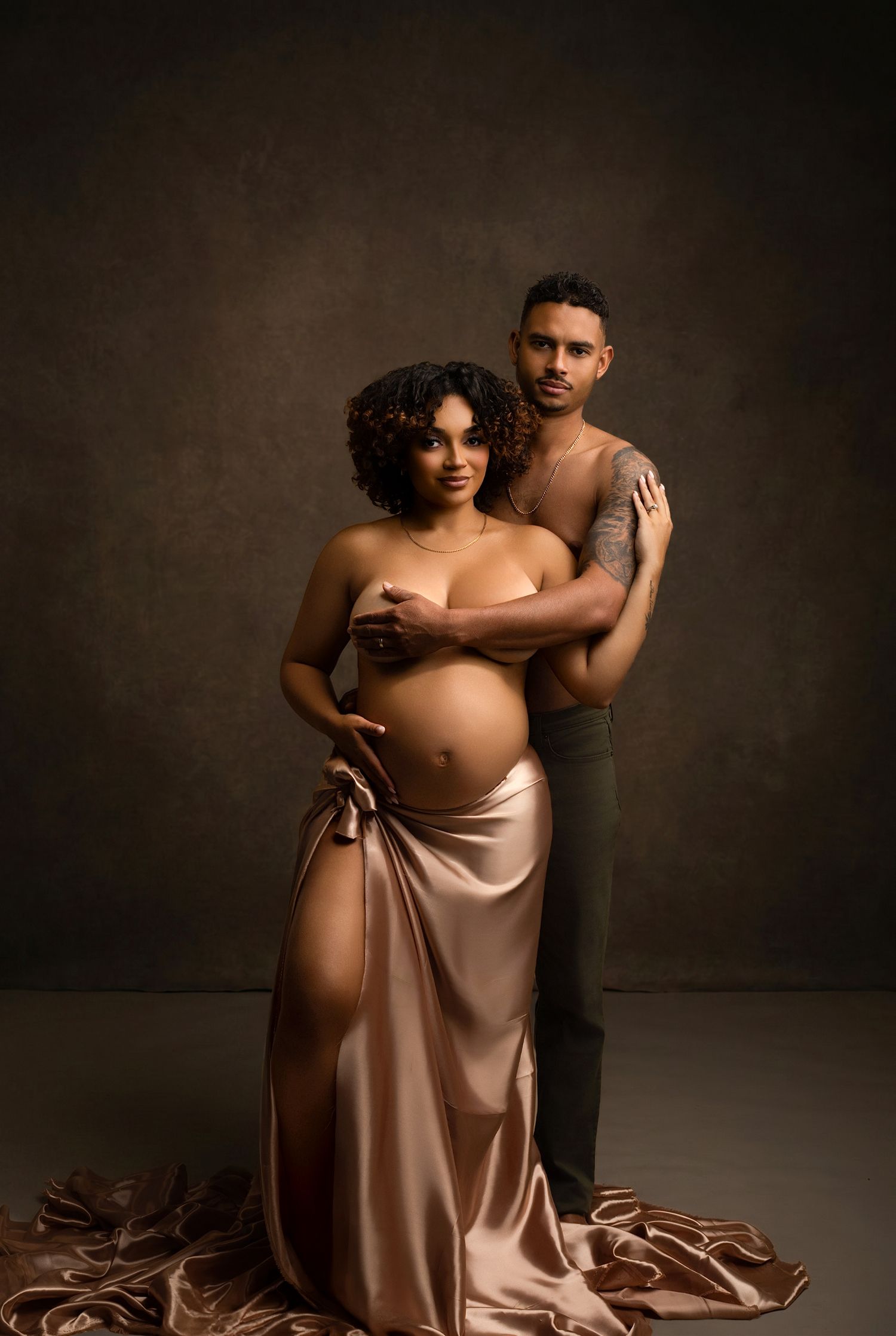 Elegant maternity photo of a pregnant woman in a satin gown embraced by her partner against a dark studio backdrop.
