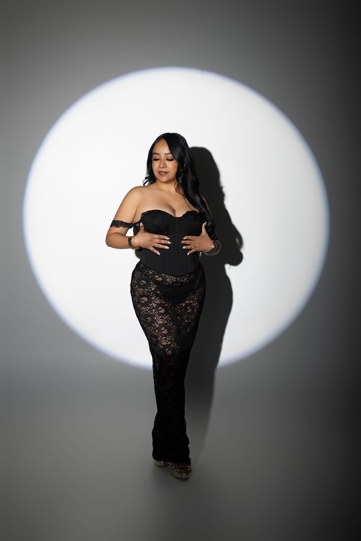 Woman in black strapless corset top and lace flare pants posing confidently in a dramatic spotlight.