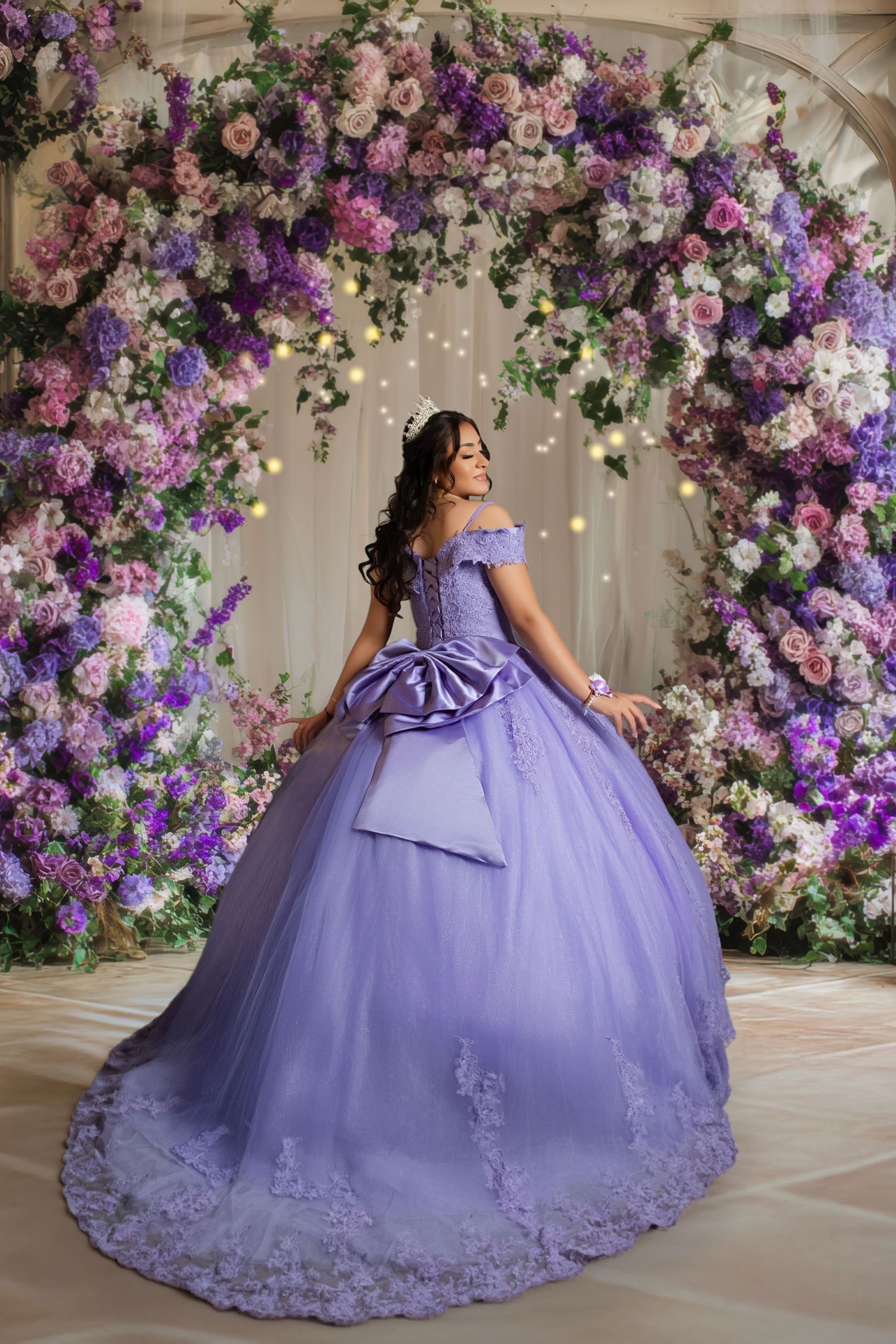 Young woman in lavender quinceañera ball gown poses before a stunning purple and pink floral arch backdrop.