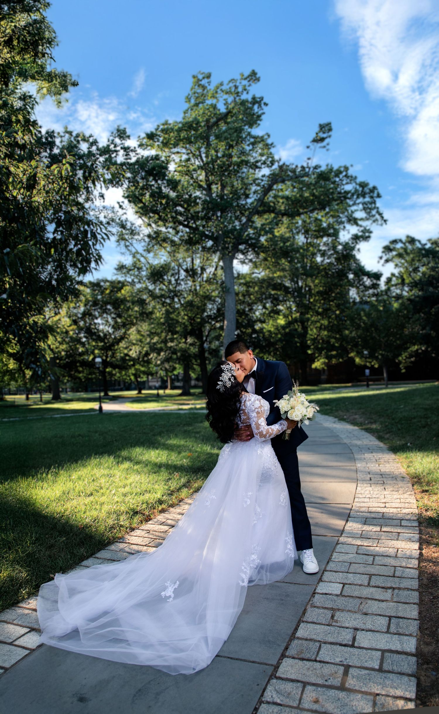 Bride and groom share a romantic kiss on a brick pathway surrounded by lush green trees on their wedding day.