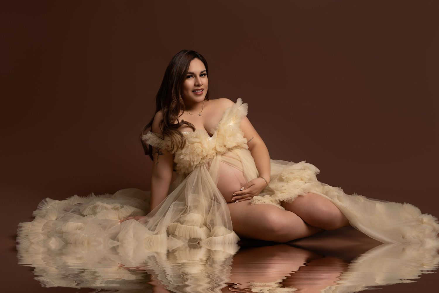 Pregnant woman in cream ruffled tulle gown poses gracefully on reflective floor against brown studio backdrop.