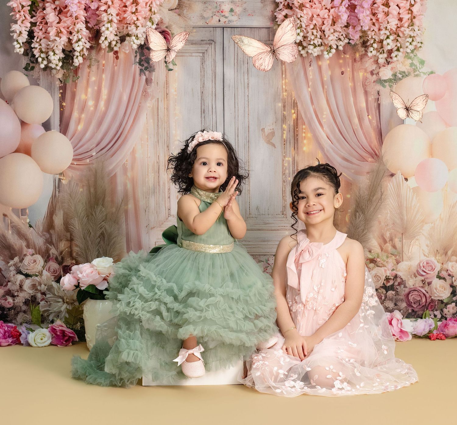 Two young girls in elegant dresses pose together at a floral spring-themed photo shoot with butterflies and balloons.