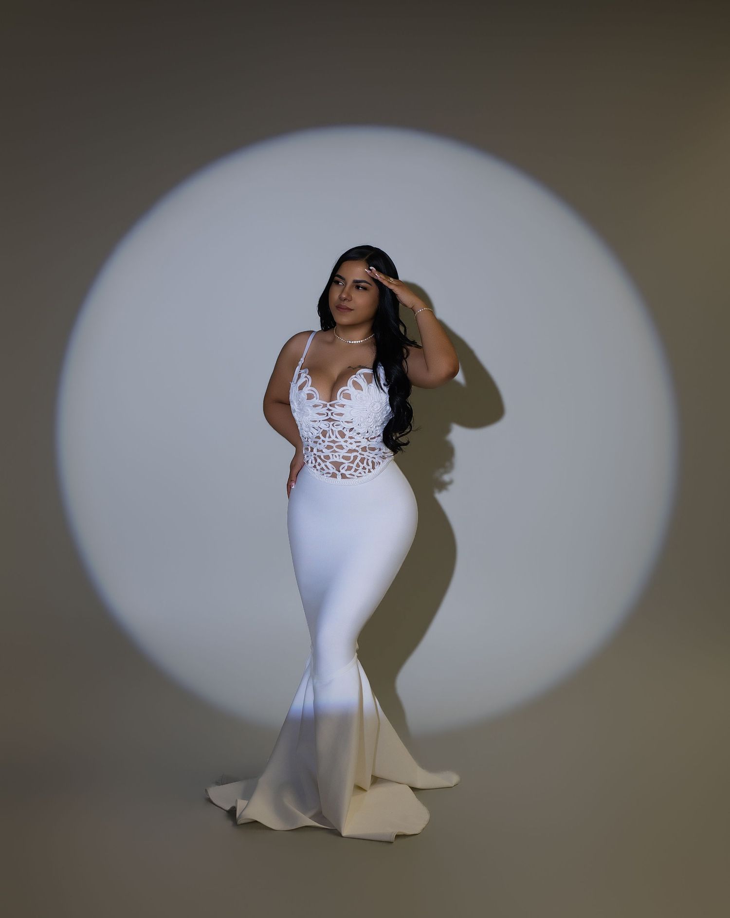 Woman in white lace mermaid gown poses elegantly in circular spotlight against neutral background.