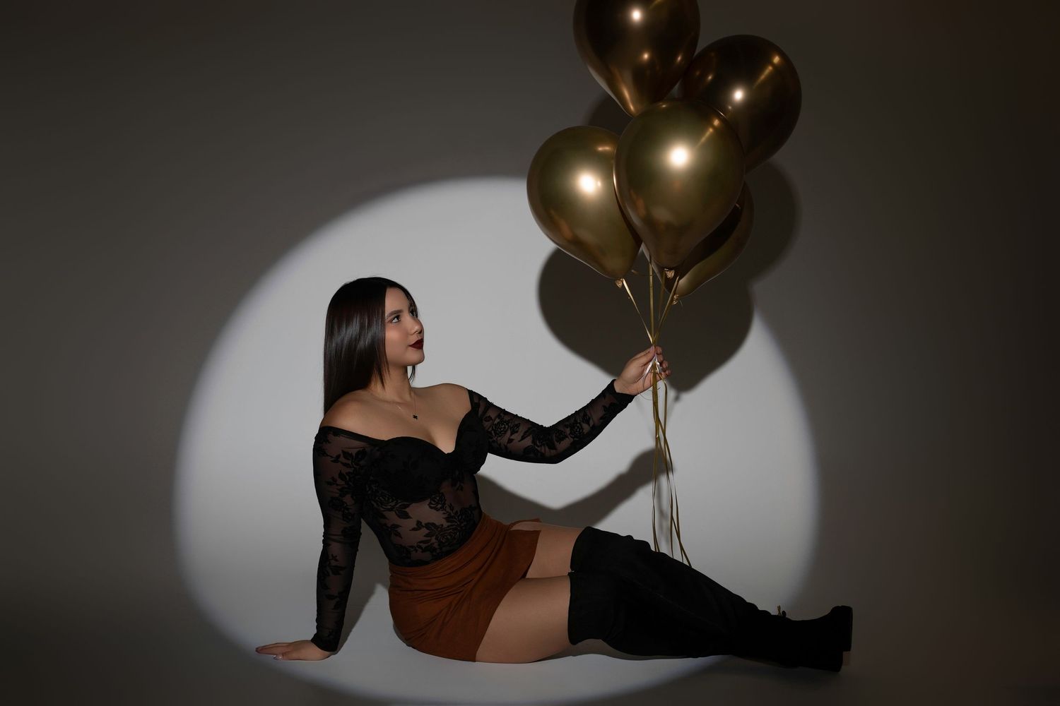 Woman in black lace top and brown skirt sitting on floor holding gold metallic balloons in dramatic spotlight.