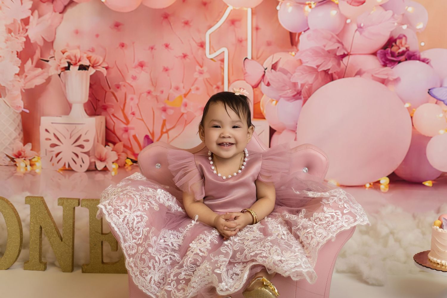 Baby girl in pink lace dress smiles at her first birthday party with balloon decorations and number one backdrop.