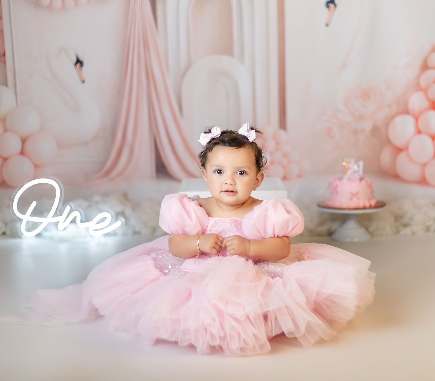Baby girl in pink tutu dress sits before first birthday cake smash setup with balloons and swan decor.