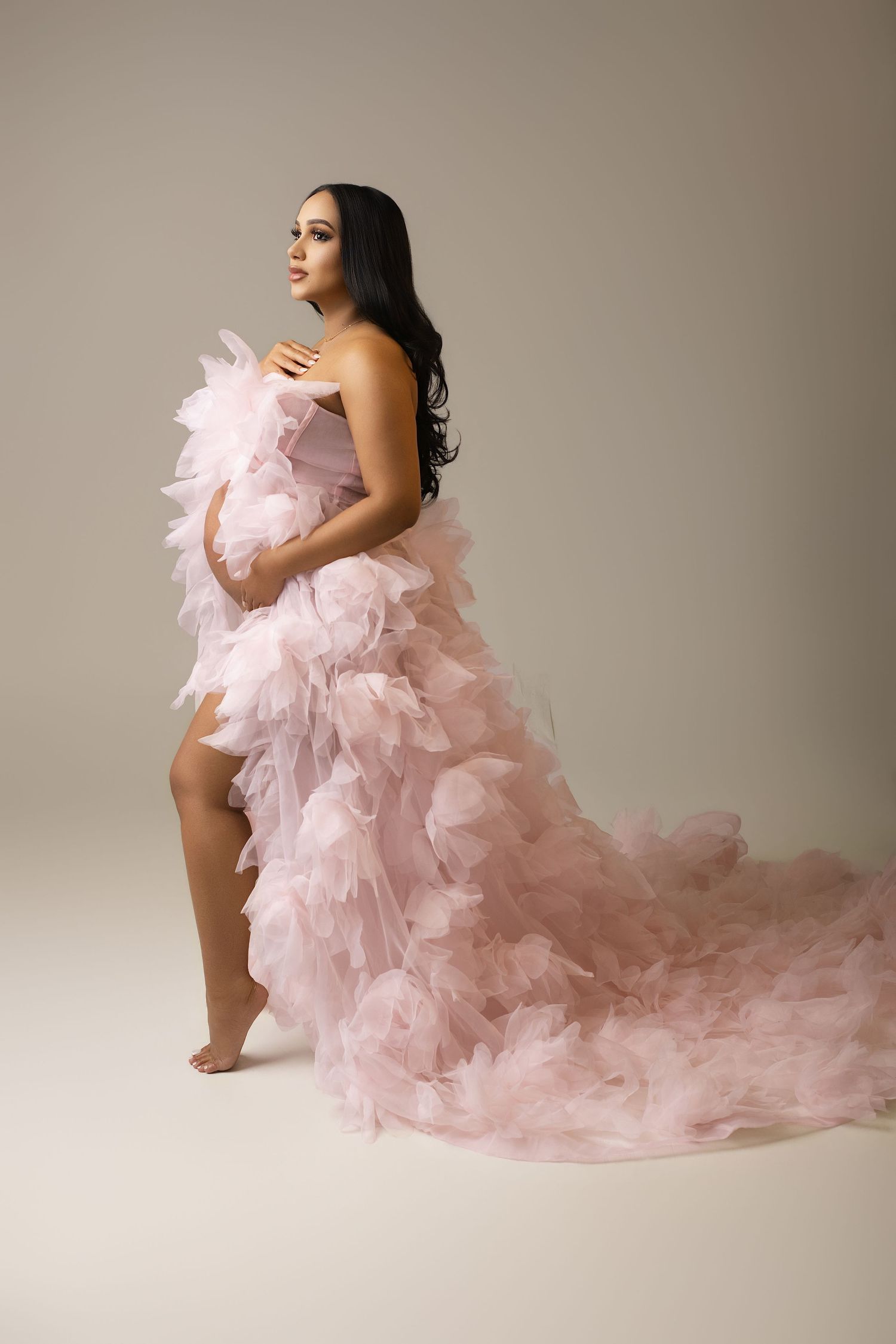Pregnant woman in a flowing pink ruffled tulle gown posing gracefully in a studio maternity photoshoot.