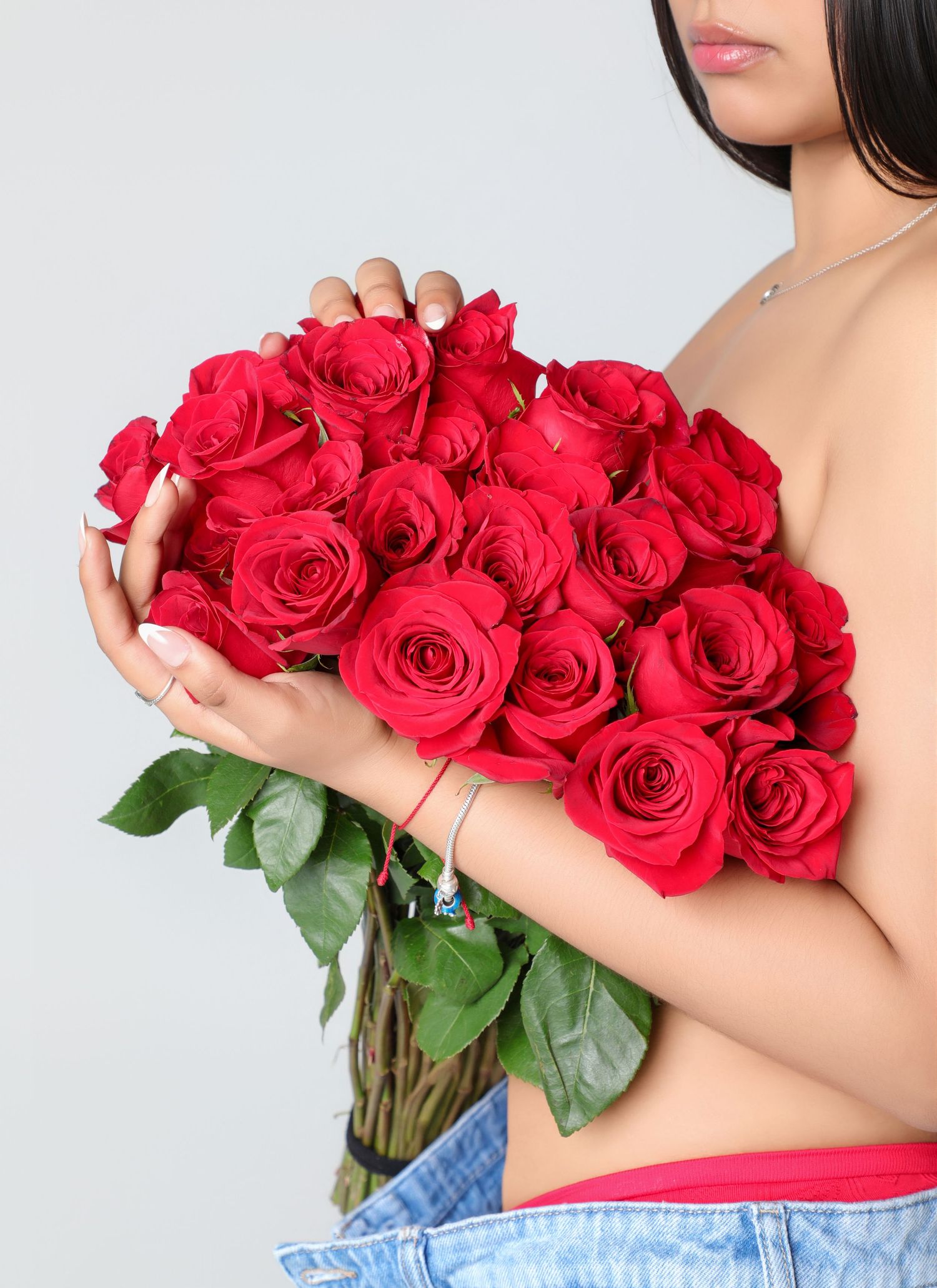 Woman holding a large bouquet of vibrant red roses with green leaves against a light gray background.