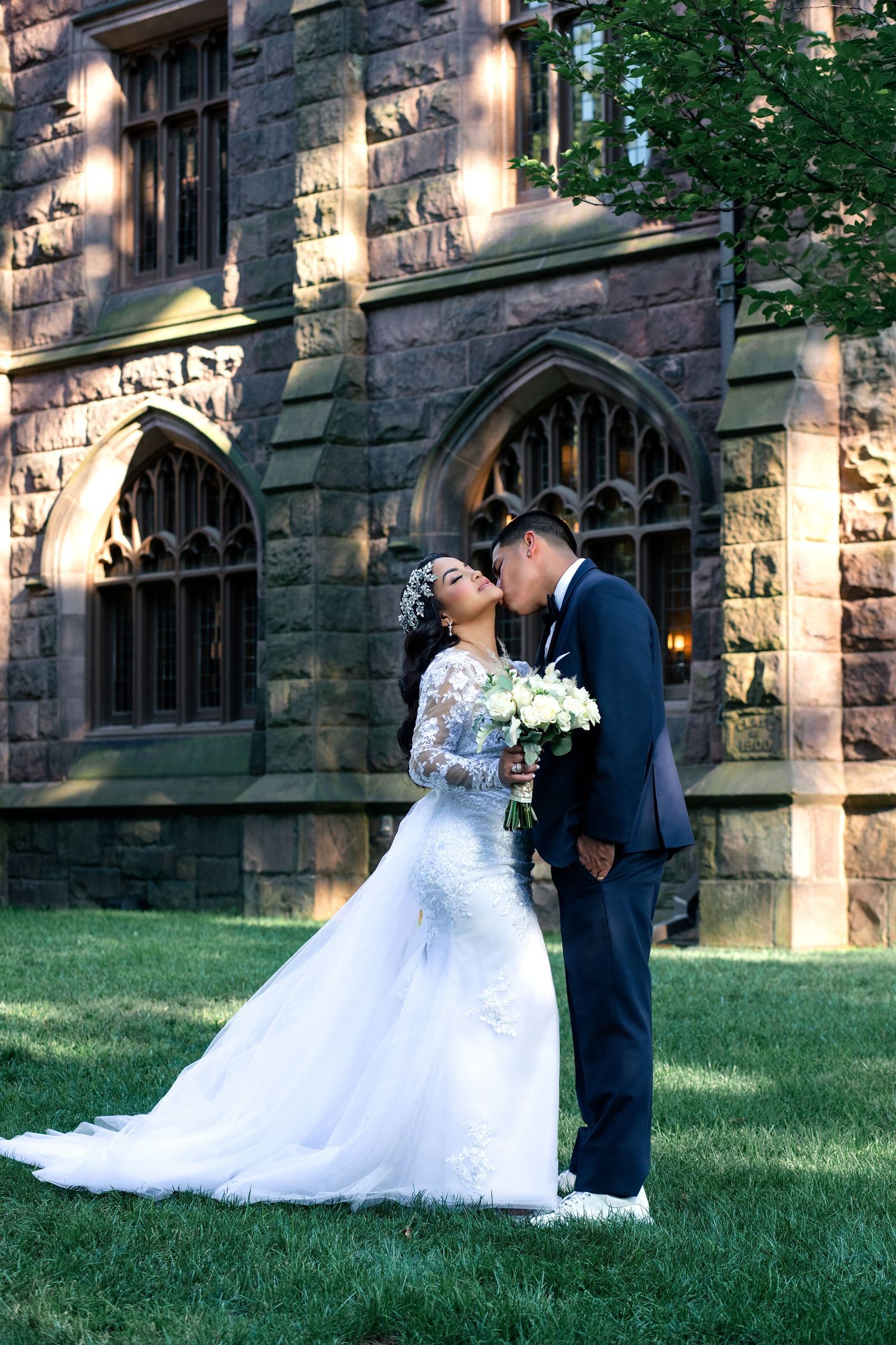 Bride and groom sharing a kiss on green lawn before a Gothic stone building, bride in lace gown holding white bouquet.