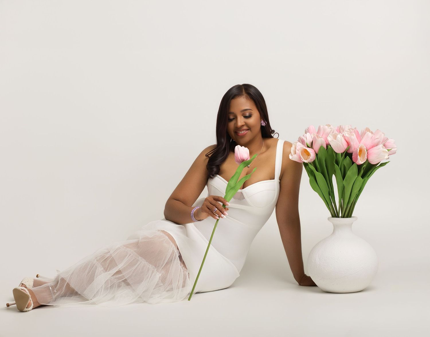 A woman in a white dress sits gracefully beside a vase of pink tulips, holding a single white rose.