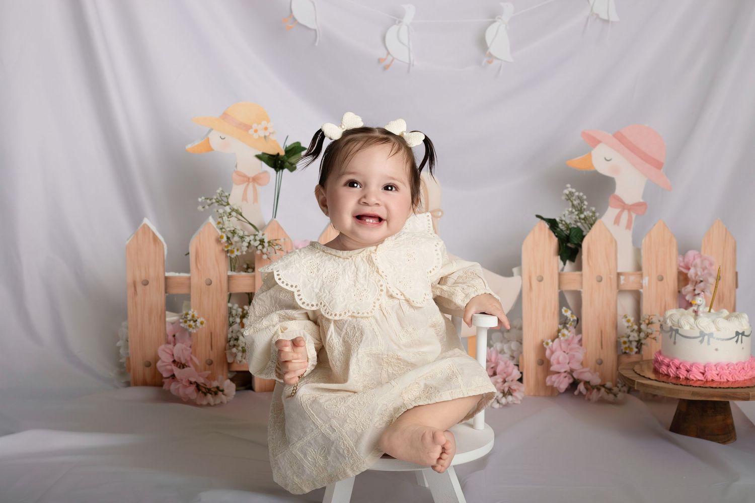 Baby girl in cream lace dress sits smiling at cake smash photo session with pastel floral fence backdrop.