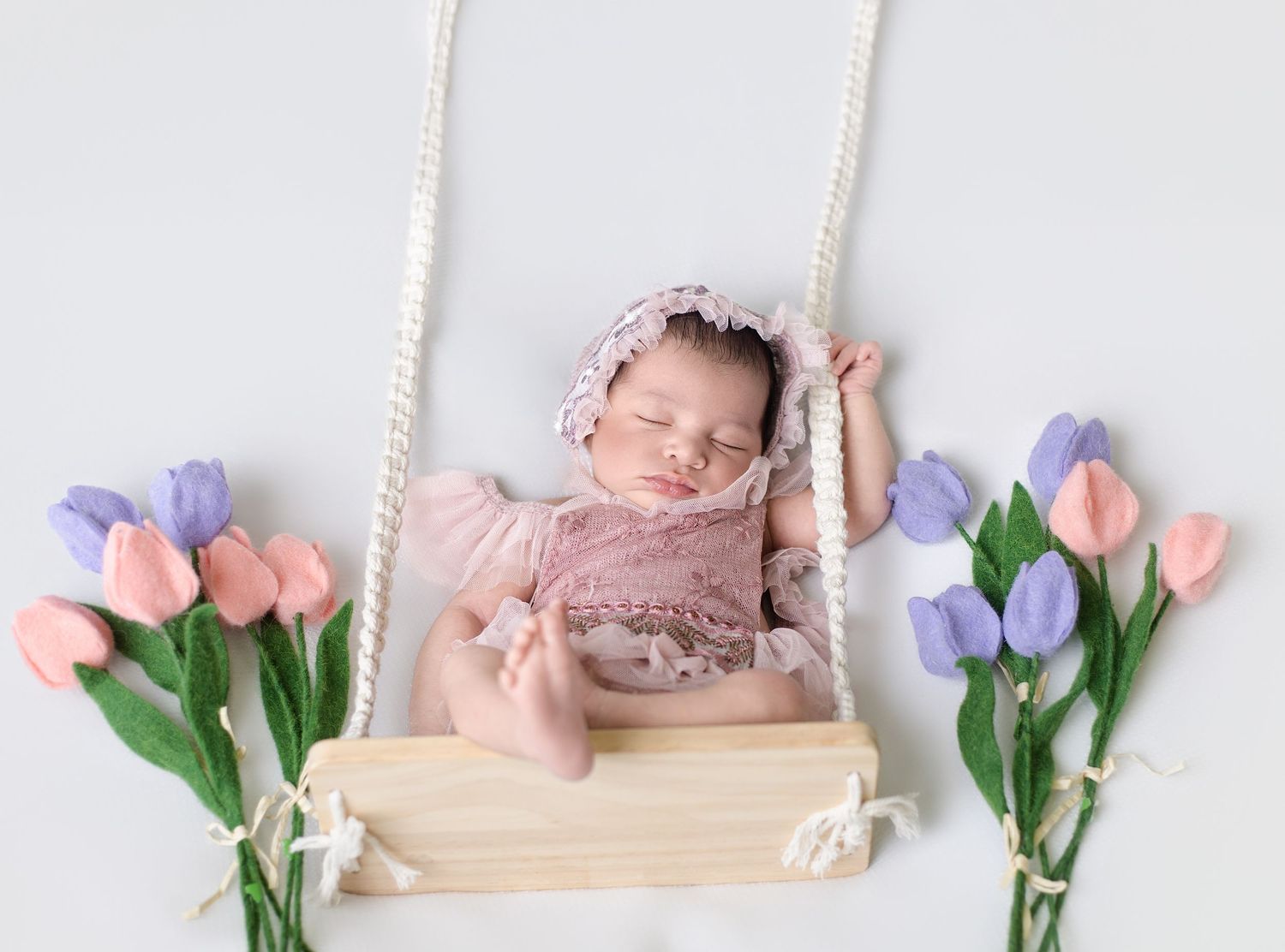 Sleeping newborn baby in pink outfit and bonnet on wooden swing surrounded by pink and purple tulips.