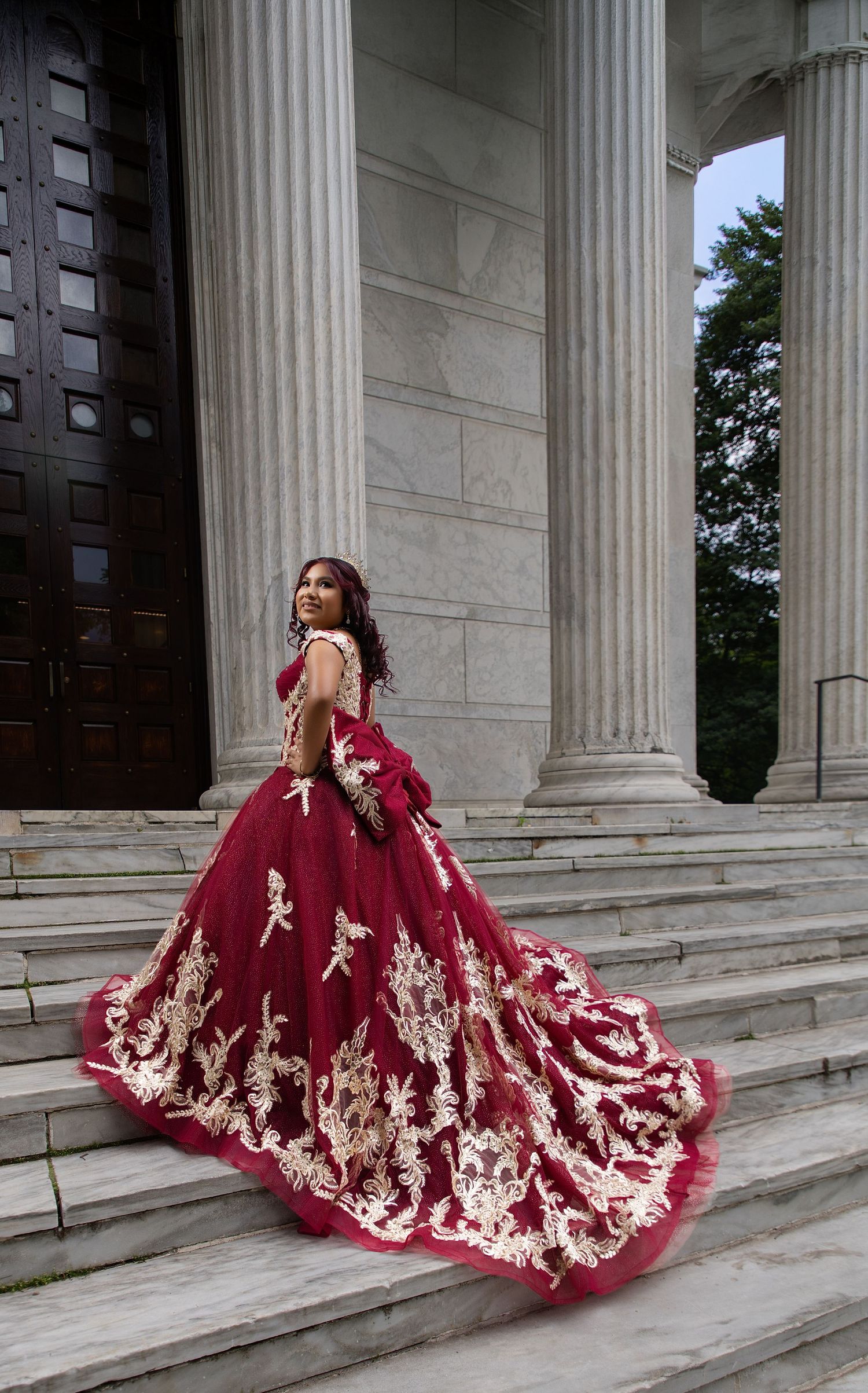 Young woman in a stunning burgundy ball gown with gold lace appliqués poses on marble steps by grand columns.