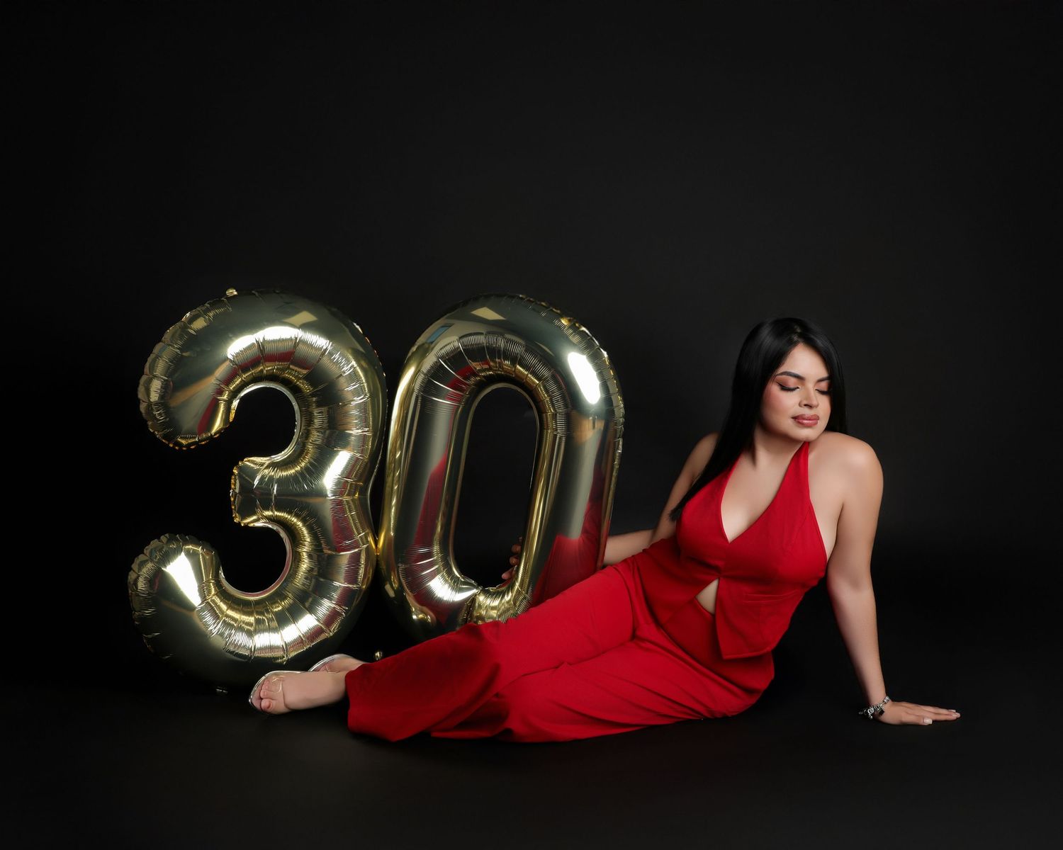 Woman in red dress posing with gold number 30 balloons on black background, celebrating her 30th birthday.