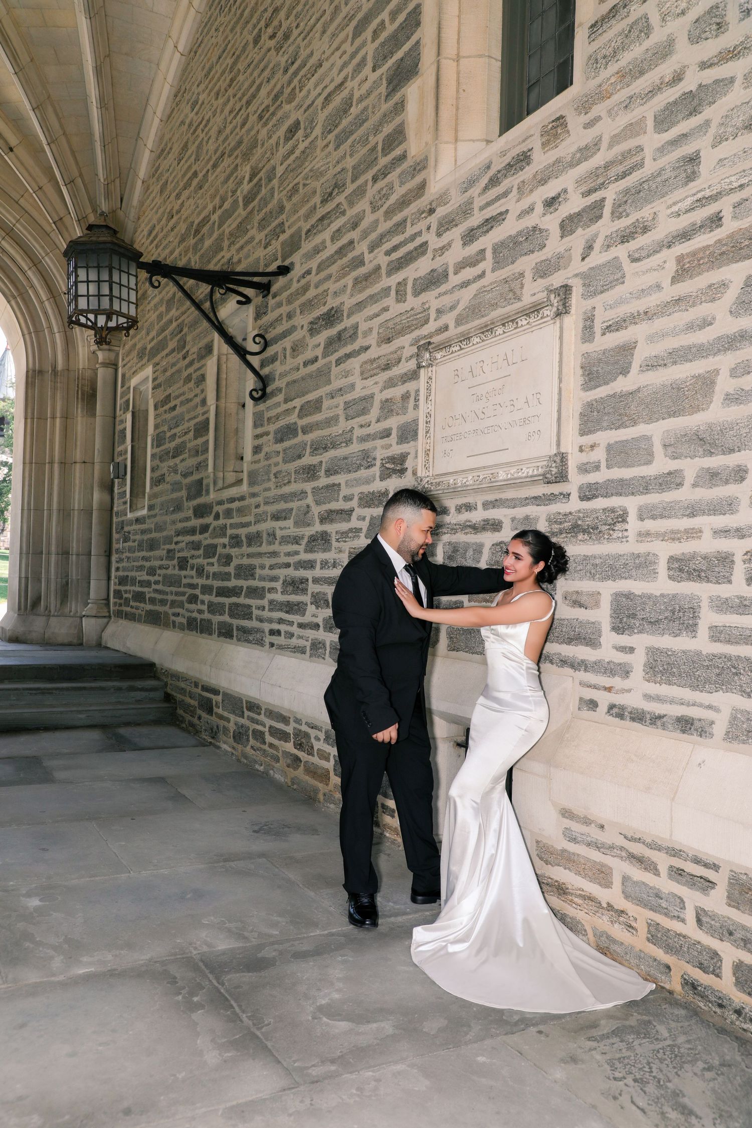 Bride in white mermaid gown and groom in black tuxedo pose romantically against a stone building wall.