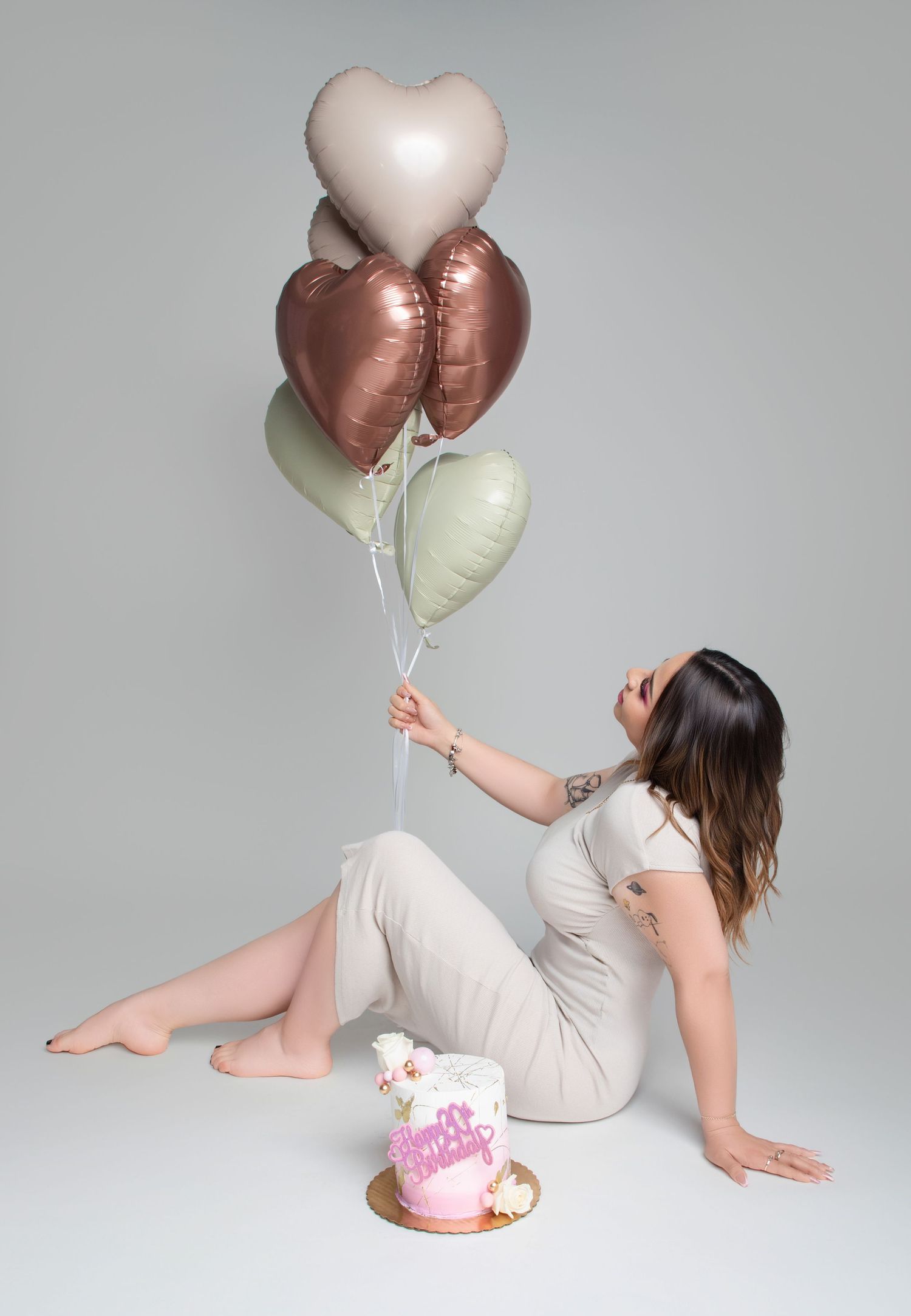 A woman sits on the floor holding heart-shaped balloons beside a birthday cake.