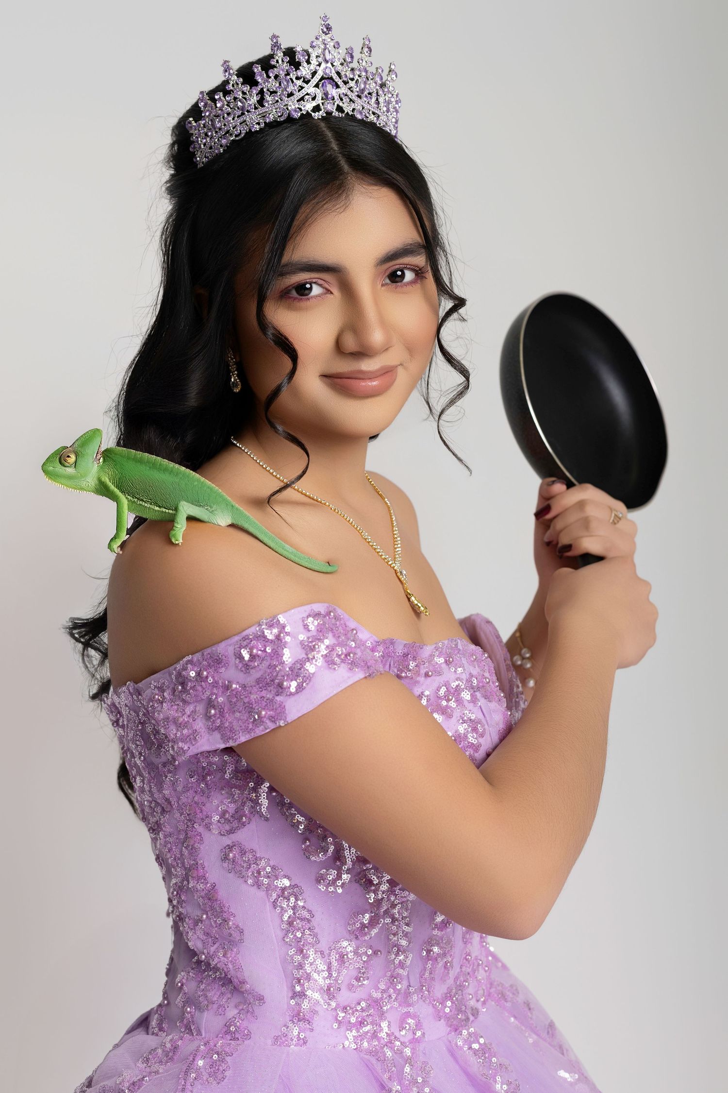 Young woman in purple quinceañera gown and tiara holds a frying pan with a green chameleon on her shoulder.