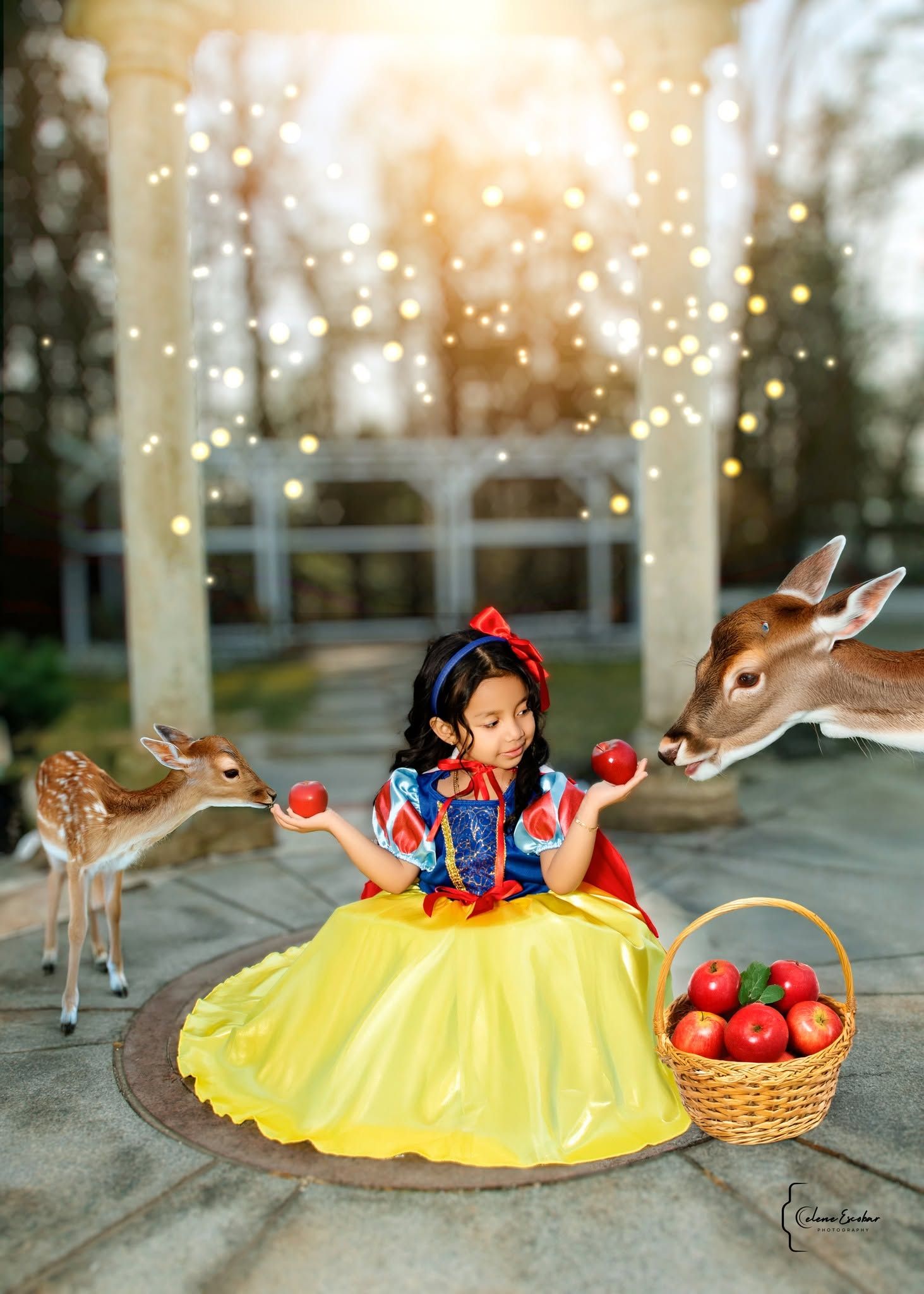Young girl in Snow White costume holds red apples while two deer approach her in a magical outdoor setting.