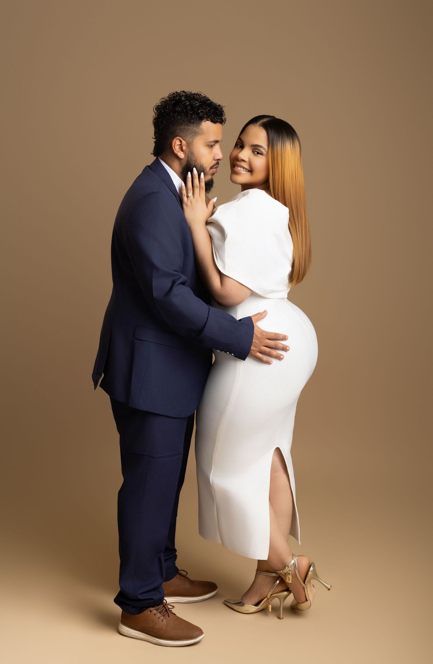 Couple posing elegantly, man in navy suit, woman in white fitted dress, studio portrait with tan background.