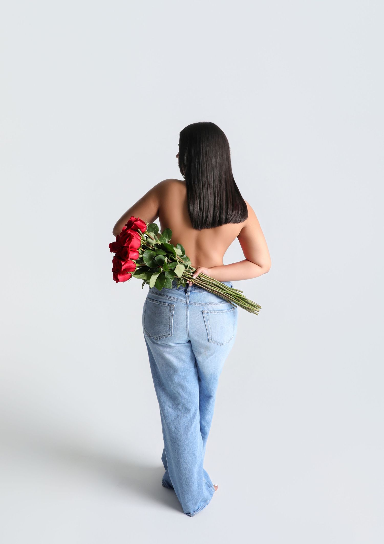 Woman with long dark hair holding red roses behind her back, wearing blue wide-leg jeans, studio shot.