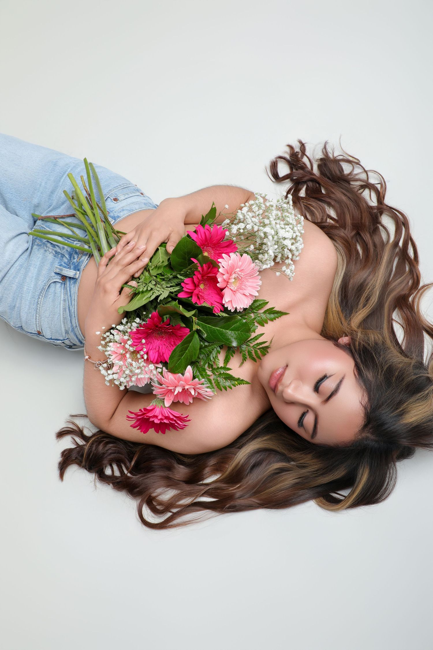 A woman with long wavy hair lies on a white background holding a bouquet of pink and white flowers, wearing blue jeans.