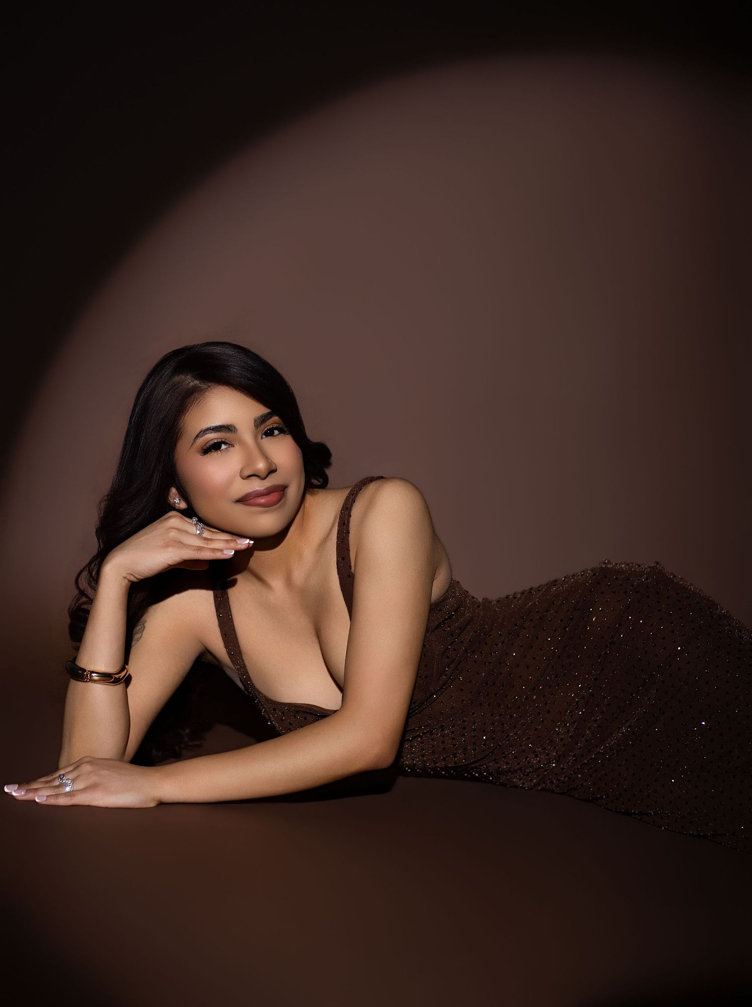 A woman in a brown sequin dress poses elegantly on the floor against a warm brown studio backdrop.