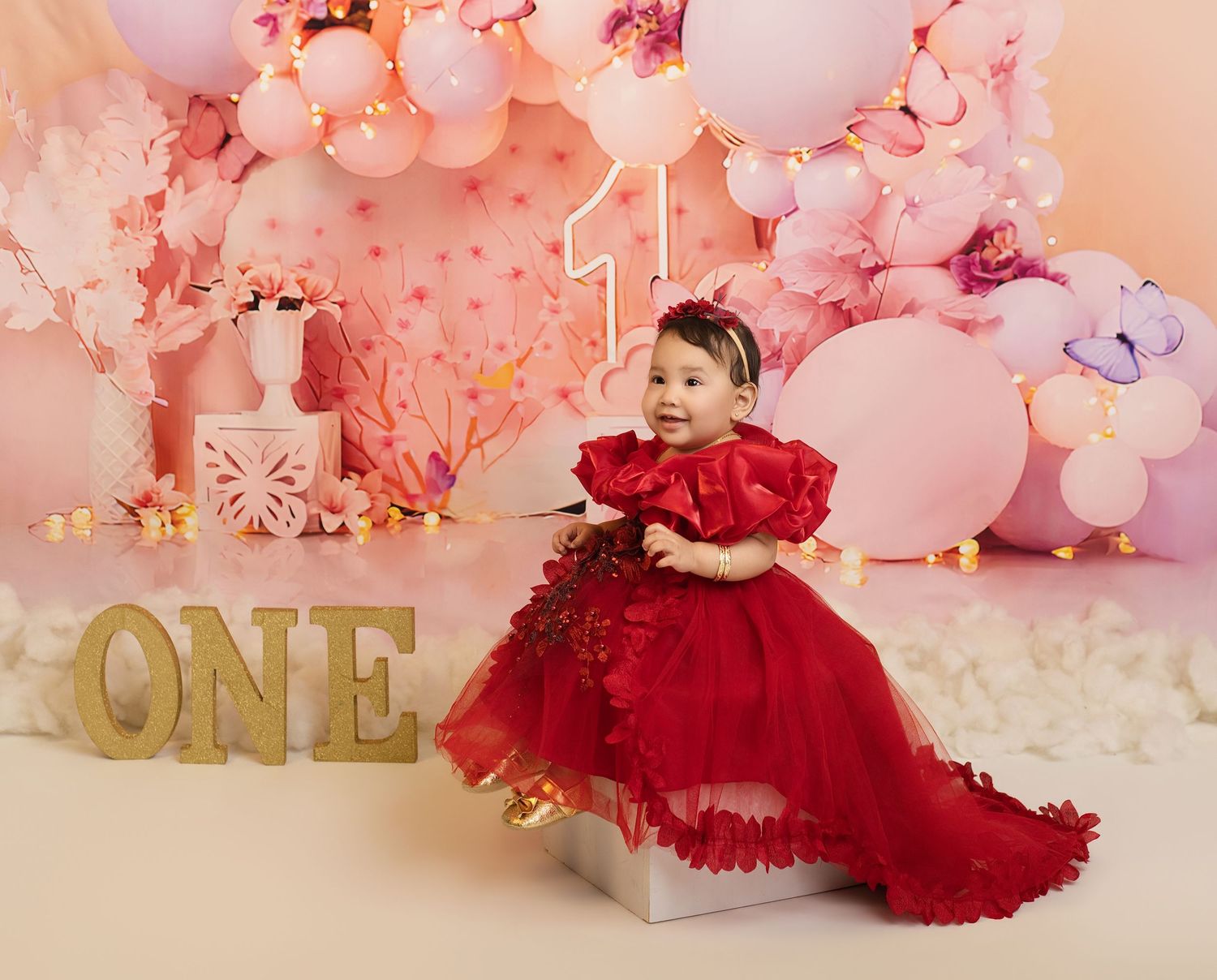 Baby girl in red dress celebrating first birthday with pink balloon arch and ONE sign.