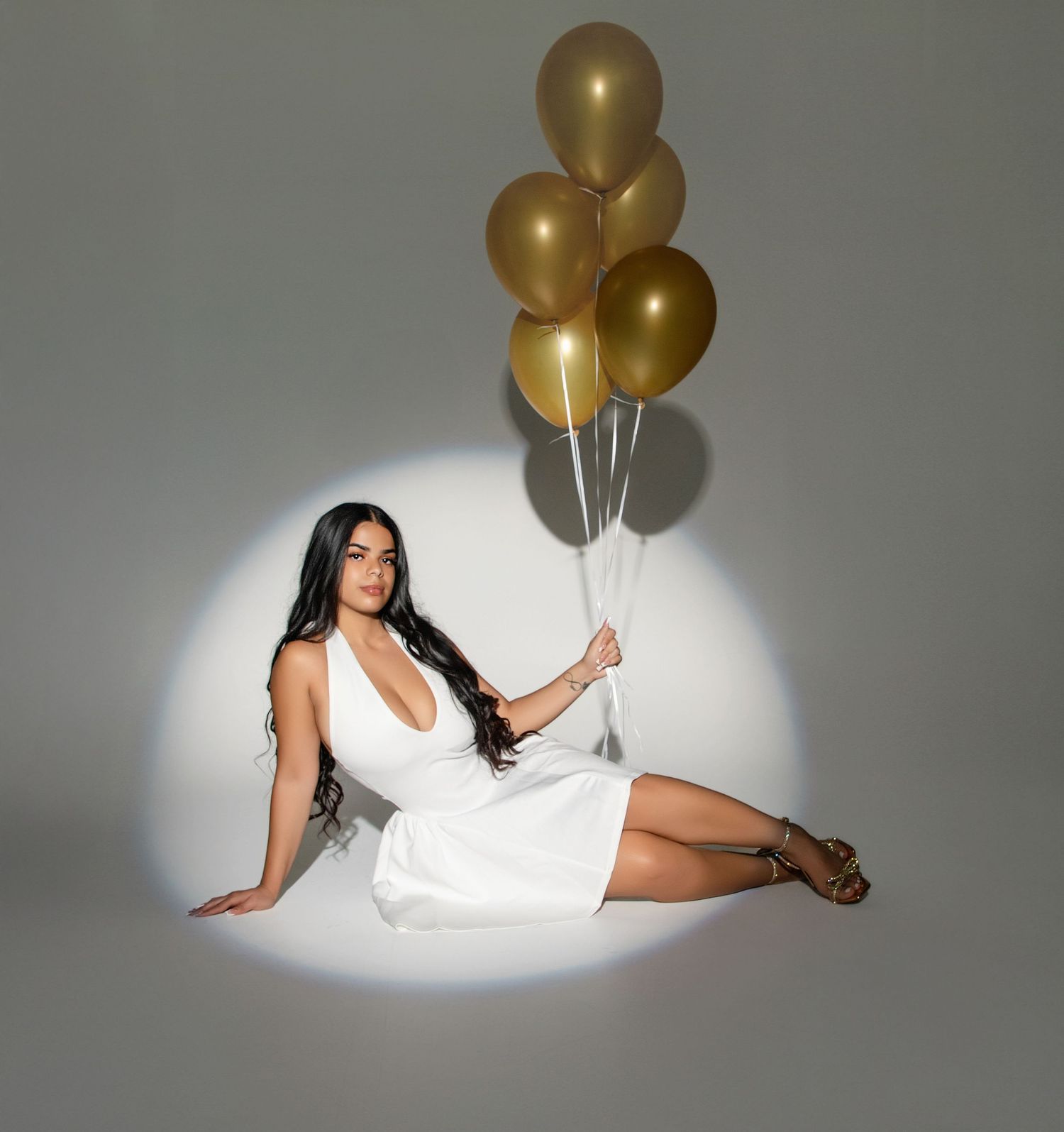 Woman in white dress sitting on floor holding gold balloons in spotlight on gray background.