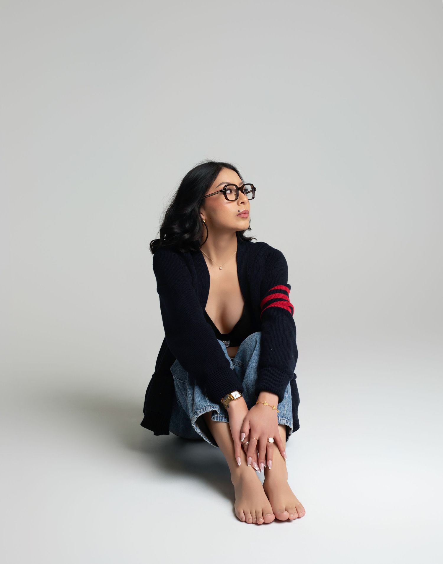 Woman in navy cardigan with red stripes and jeans sits barefoot on floor, wearing glasses, gazing upward.