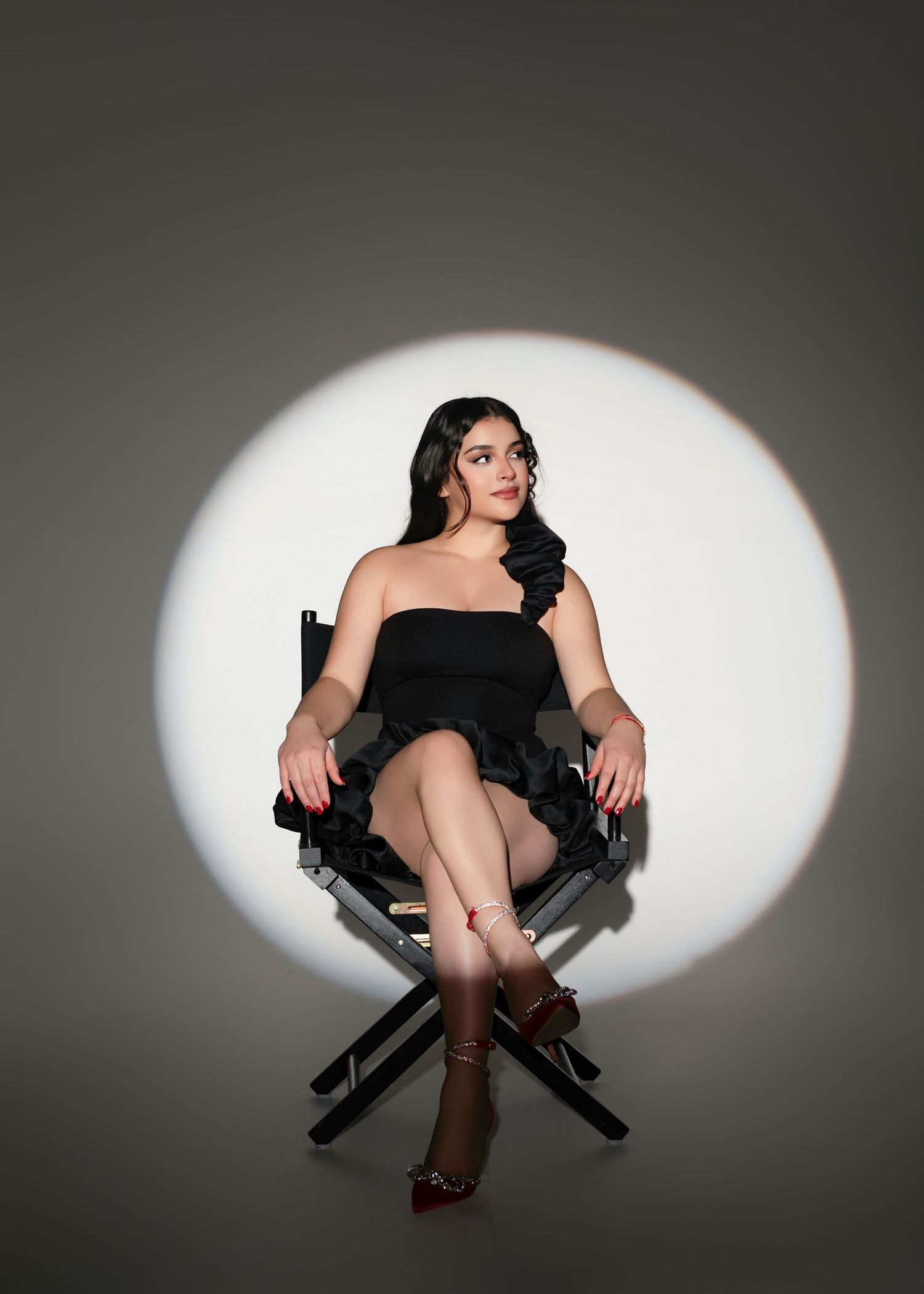Woman in black one-shoulder mini dress seated in director's chair against dramatic spotlight background.