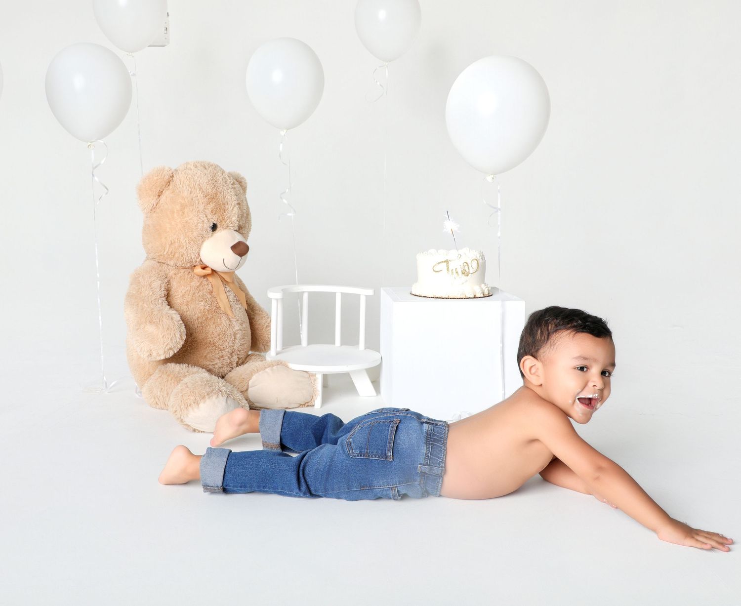 Smiling toddler boy in jeans poses with a large teddy bear and birthday cake on white balloons background.