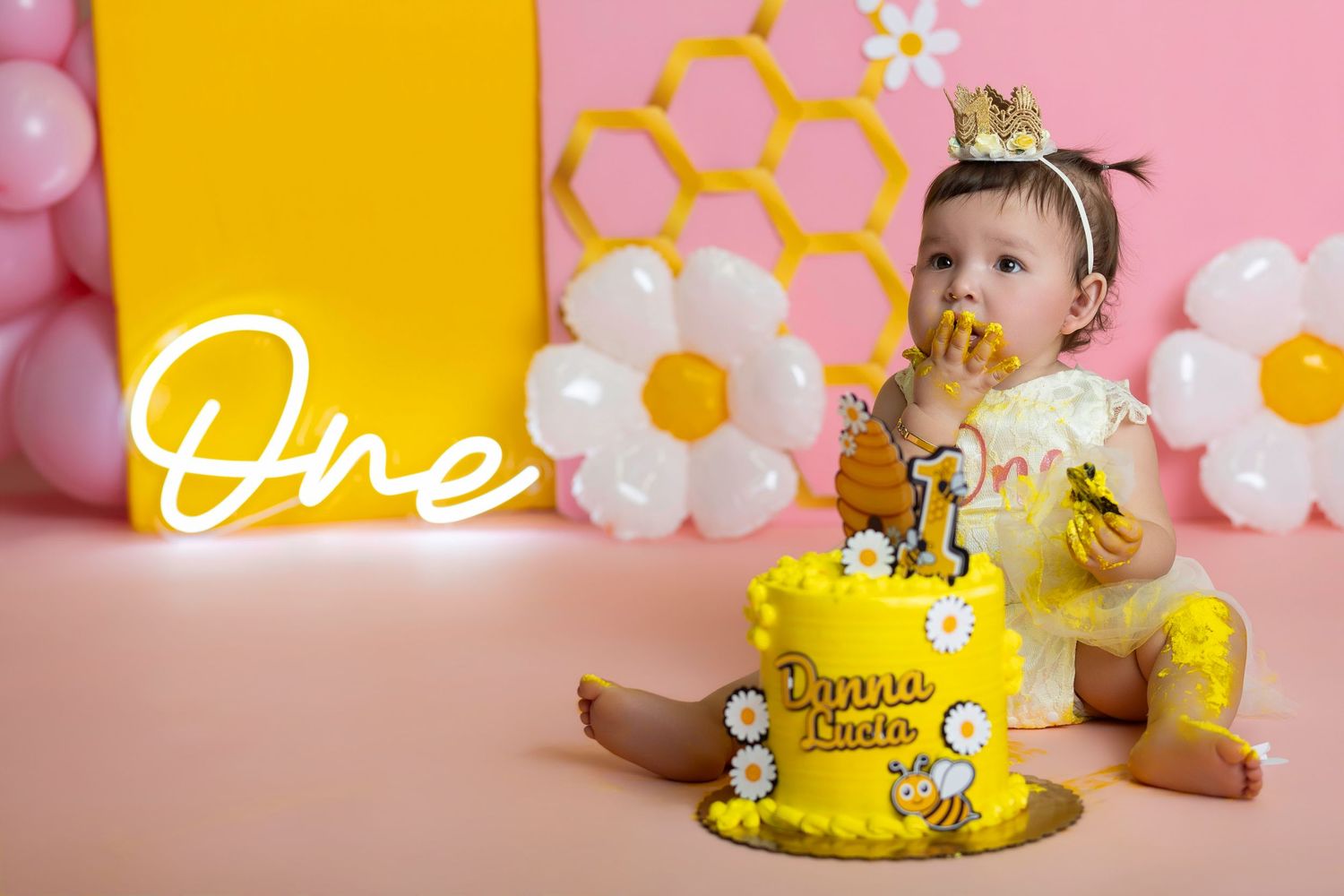 Baby girl in yellow bee outfit enjoys her first birthday cake smash with daisy balloons and pink honeycomb backdrop.