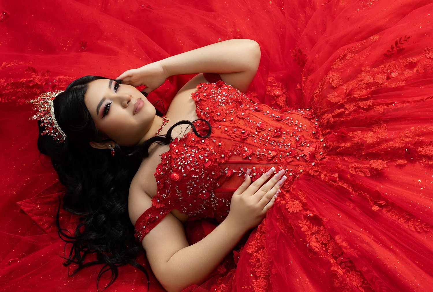 Young woman in a stunning red beaded ball gown with floral details and crown, lying elegantly on red tulle fabric.