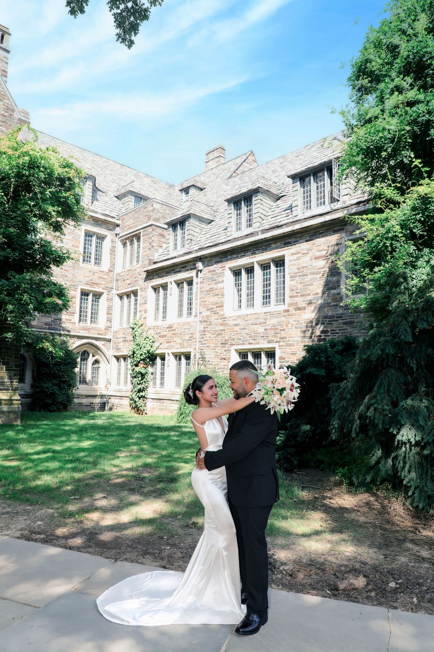 Bride and groom embrace in front of historic stone building on sunny wedding day.
