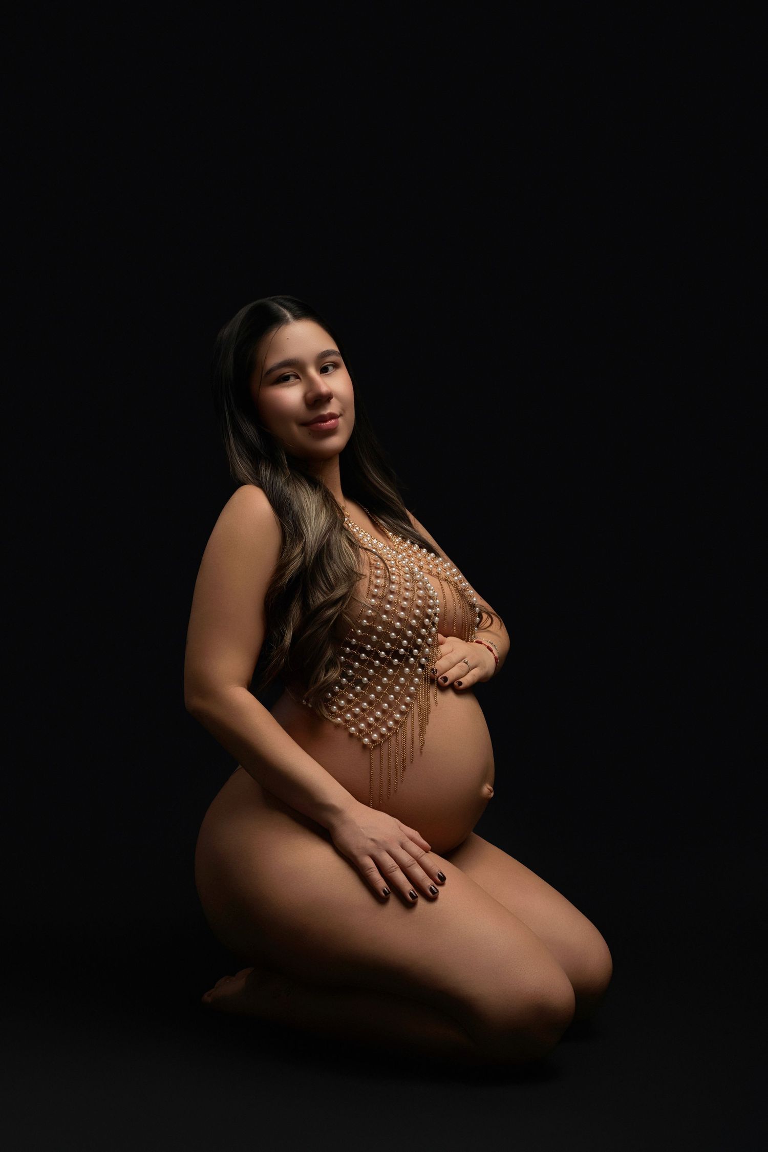 Pregnant woman in beige pearl bodysuit kneeling on studio floor against black background, cradling baby bump.