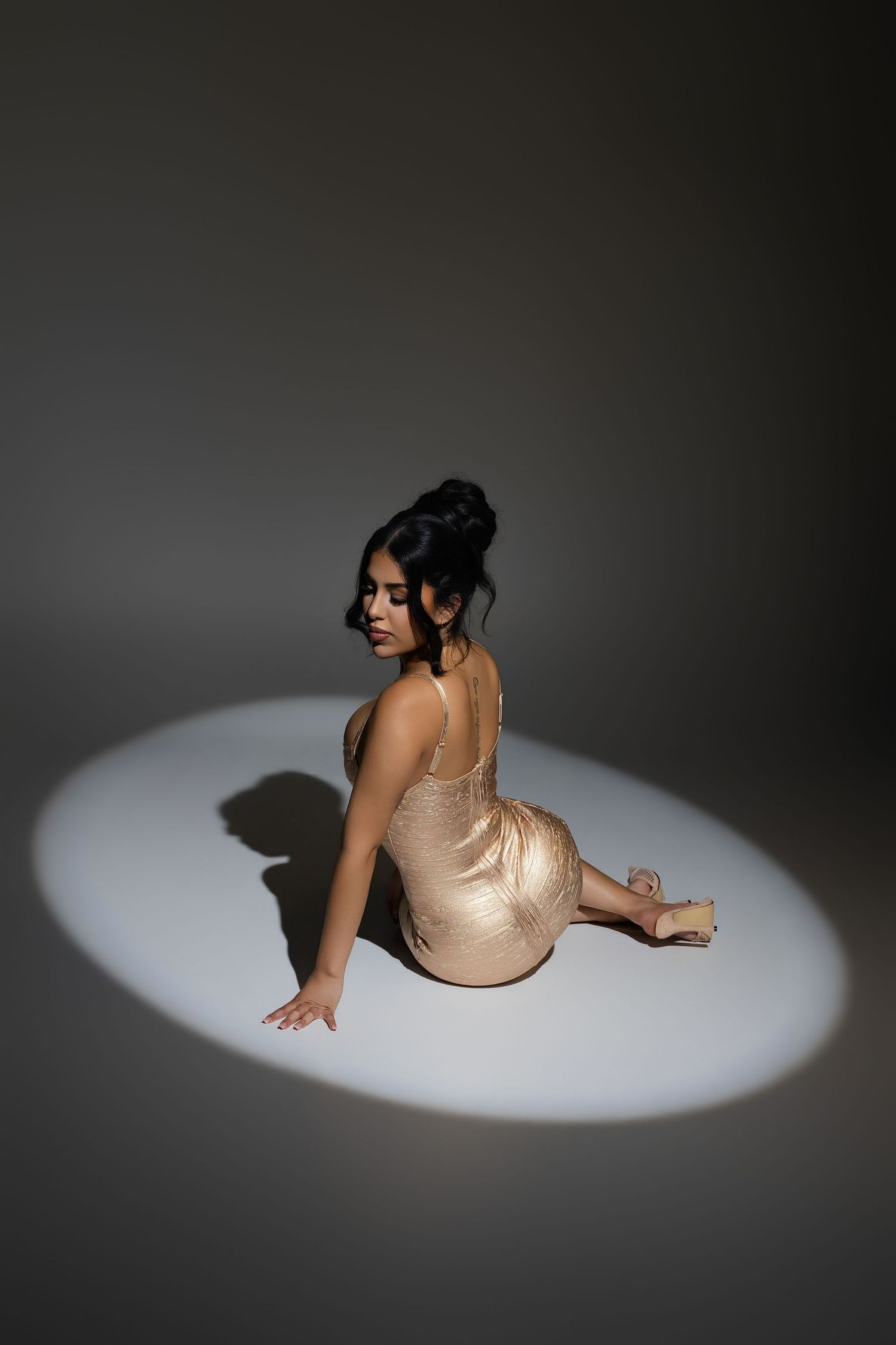 Elegant woman in beige satin dress sitting gracefully in a spotlight on dark background.