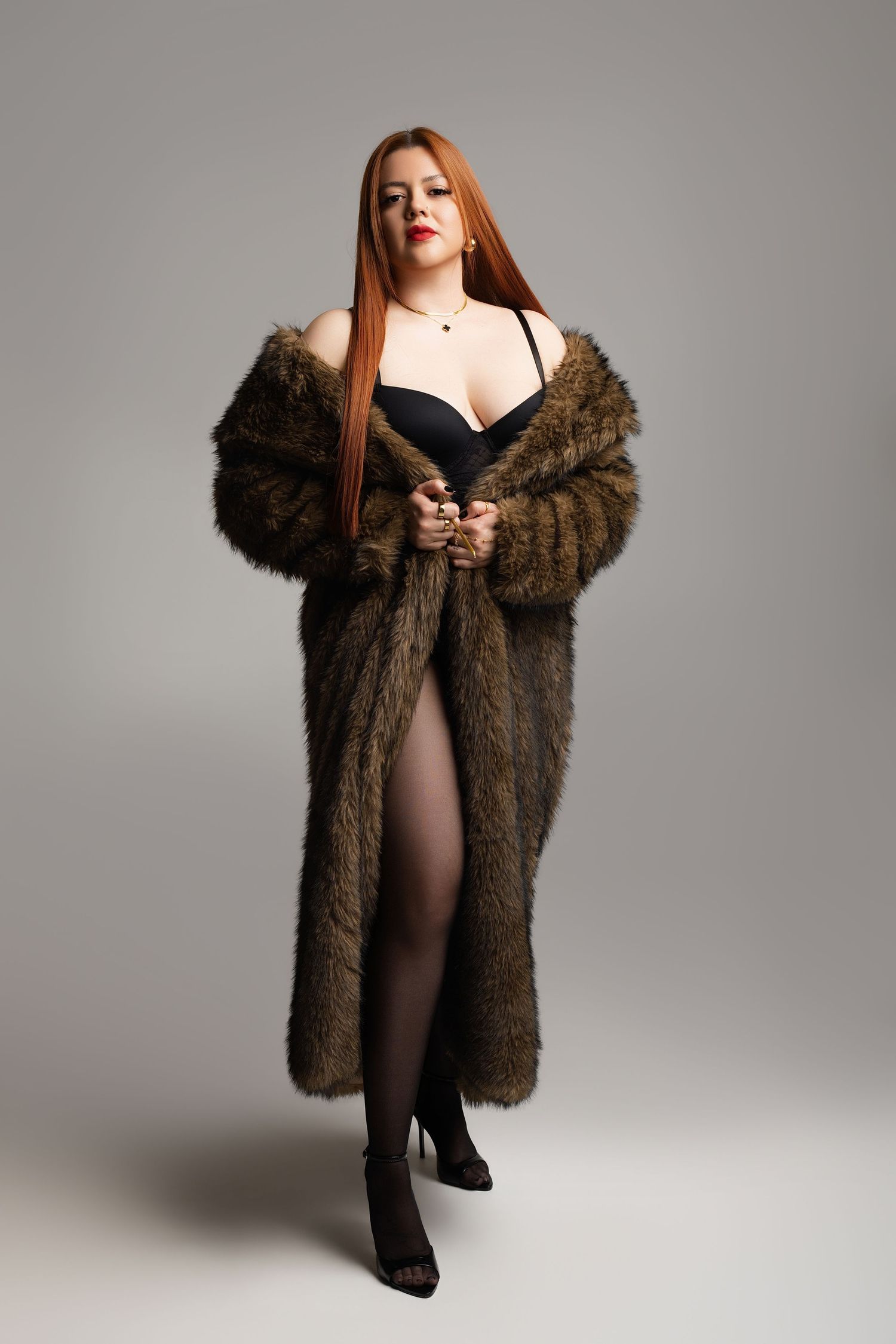 Curvy redhead woman in black lingerie and oversized brown fur coat posing confidently in studio.