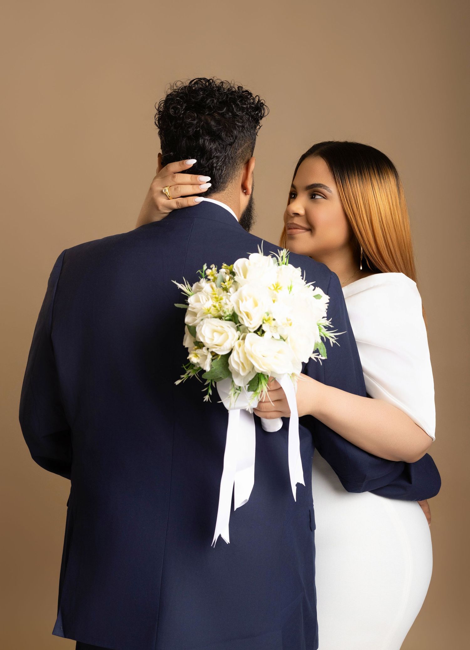 A bride holding white roses embraces a groom in a navy suit against a tan background.