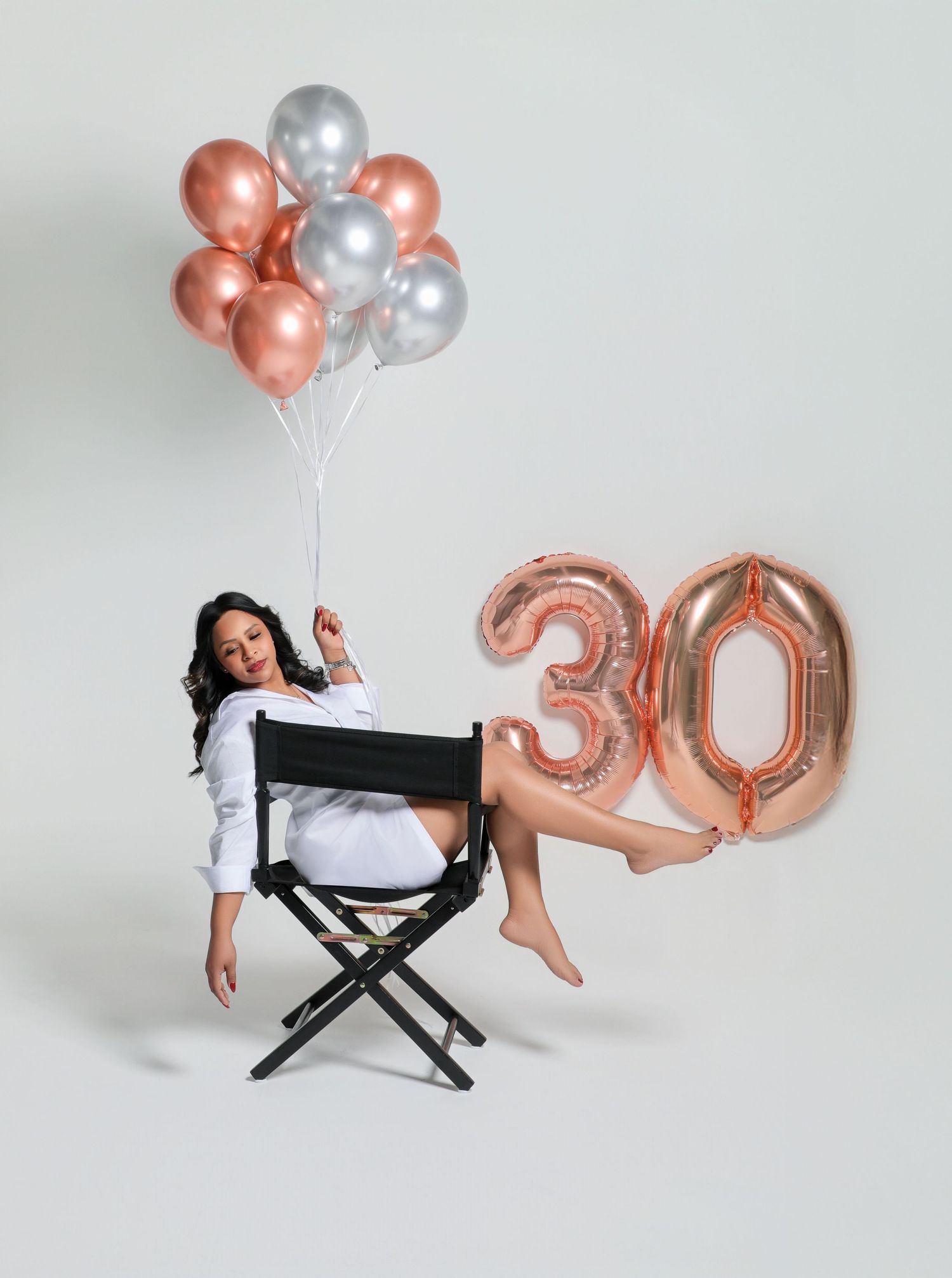 Woman celebrating 30th birthday sitting in director's chair with rose gold and silver balloons and number 30 balloon.